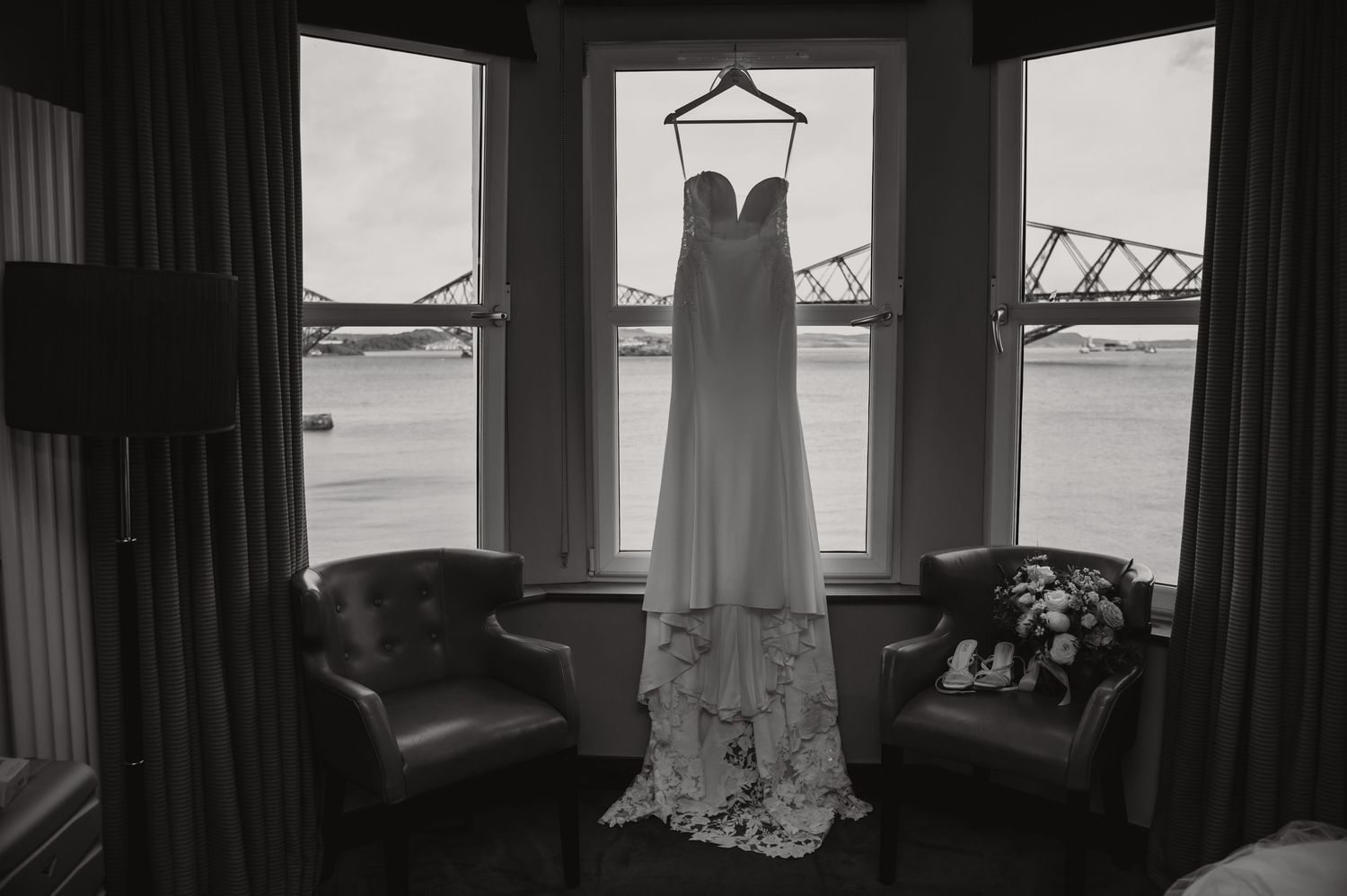A wedding dress hangs in silhouette against windows overlooking the Forth Bridge in Scotland.