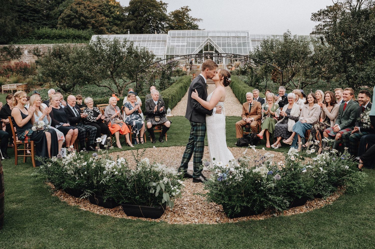 Newlyweds share first kiss in circular garden while wedding guests applaud.