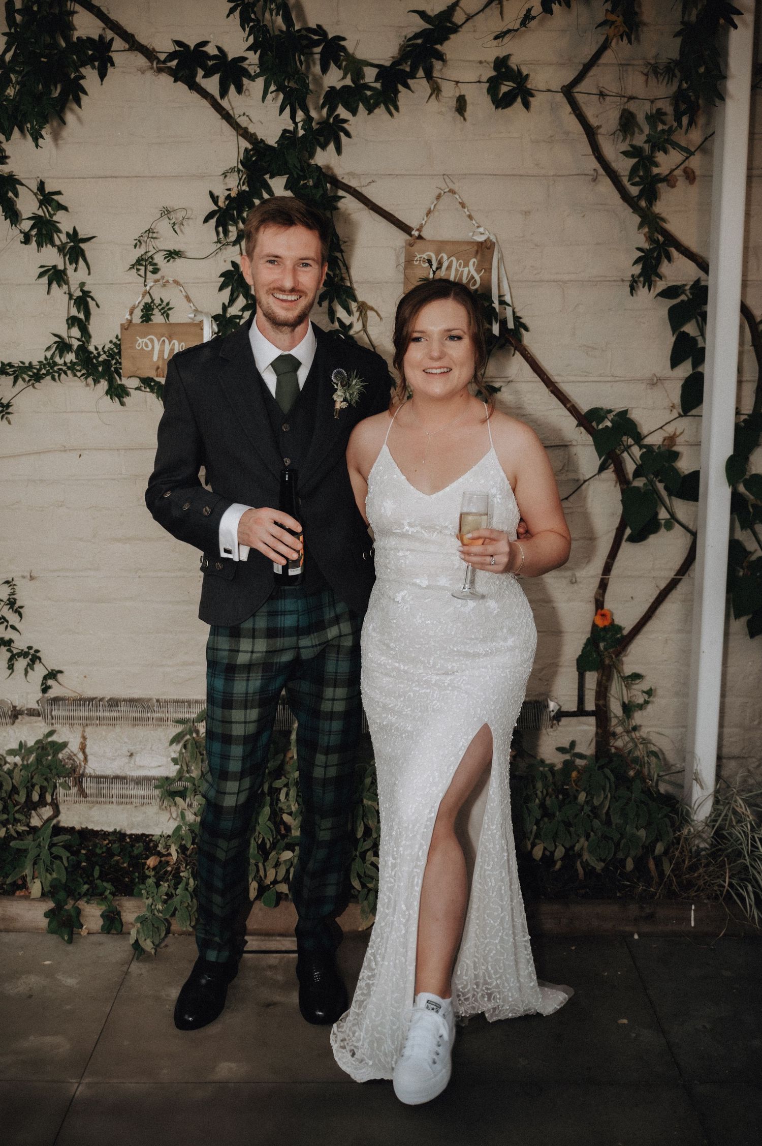 Newlyweds pose with drinks, groom wearing traditional Scottish kilt and bride in elegant white dress.