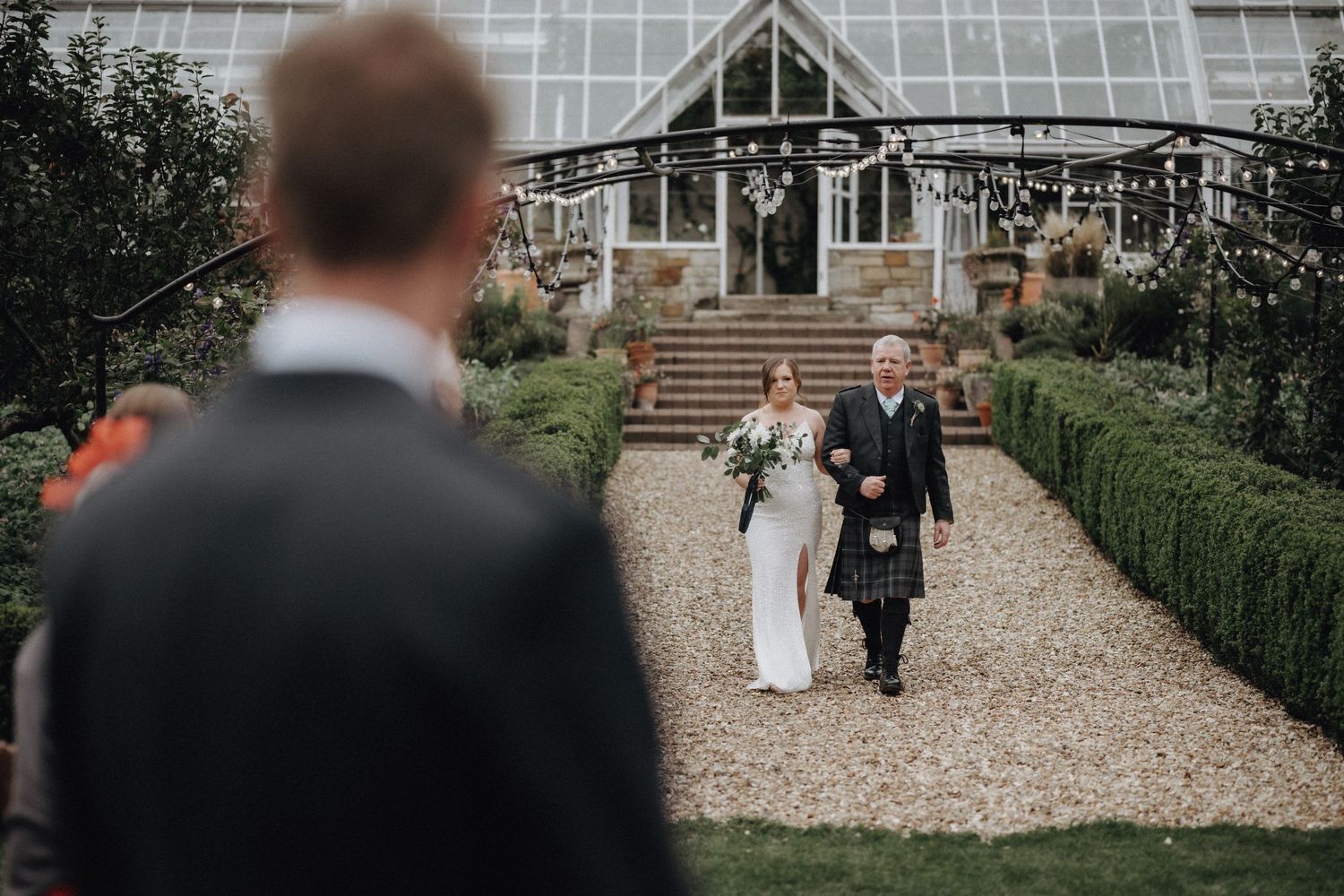 Wedding couple exits greenhouse venue on gravel path while guests look on.