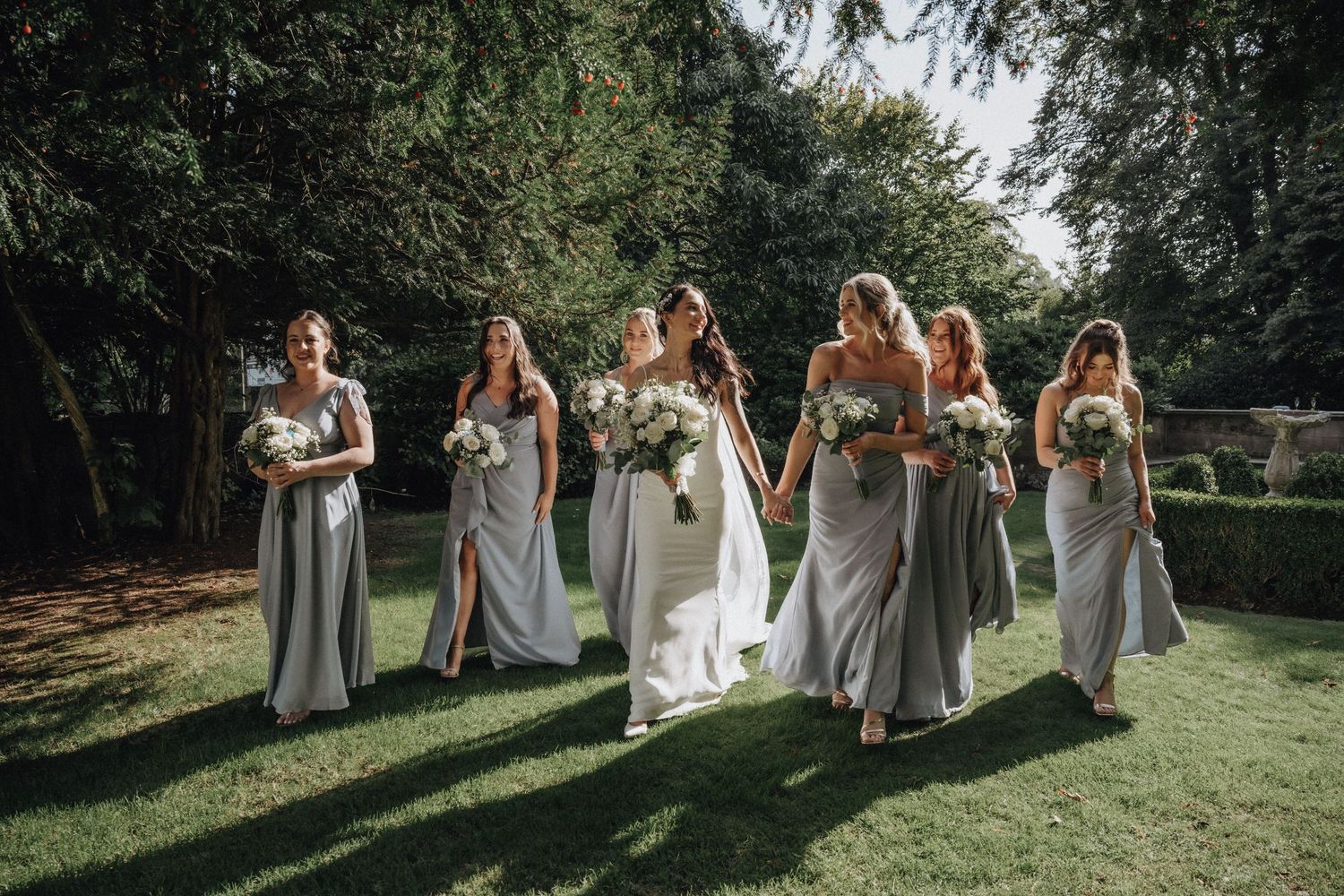 Bridal party in soft gray dresses walking across sunny garden lawn during wedding celebrations.