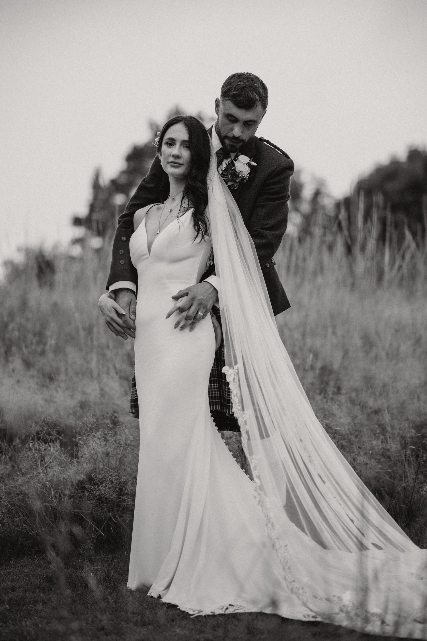 Black and white portrait of bride and groom embracing in a grassy field with long flowing veil.
