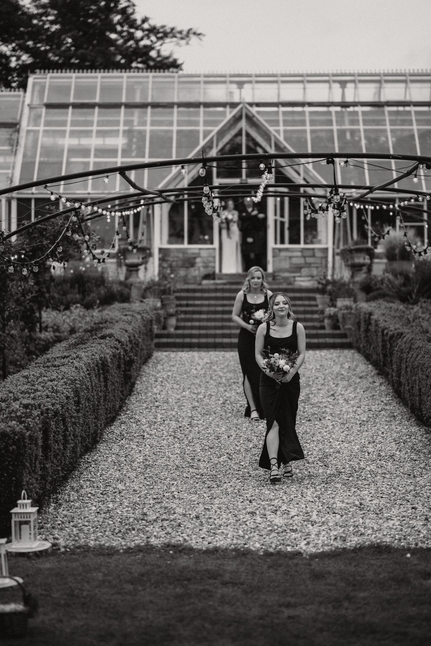 Two figures stand on a gravel path leading to a greenhouse decorated with string lights and lanterns.