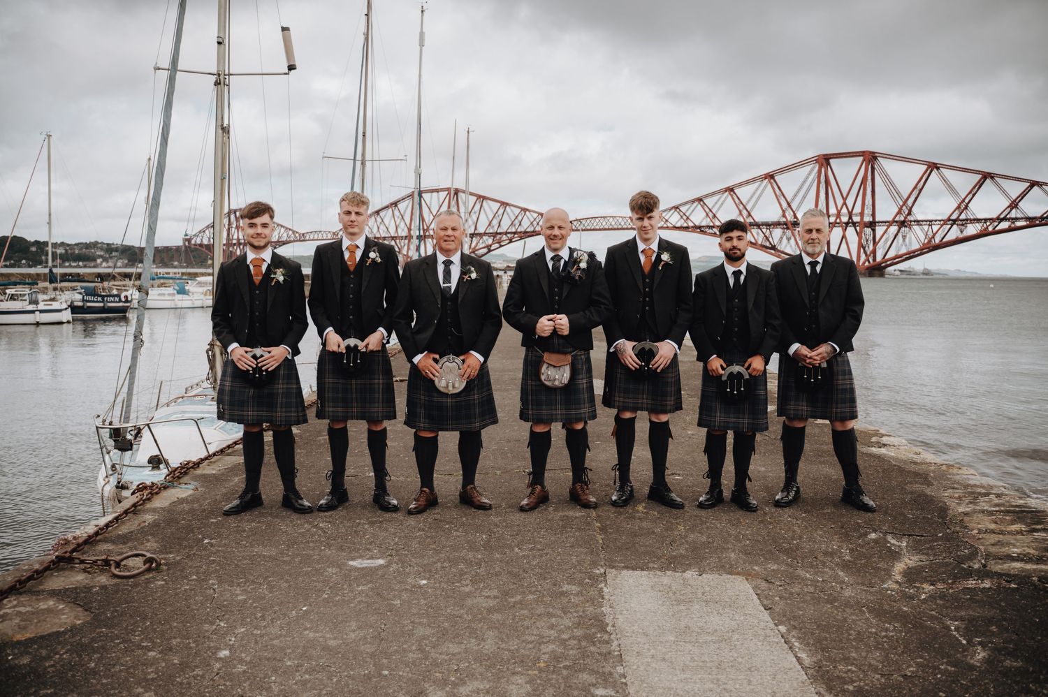 Wedding party in traditional Scottish kilts posing by harbor with iconic Forth Bridge.