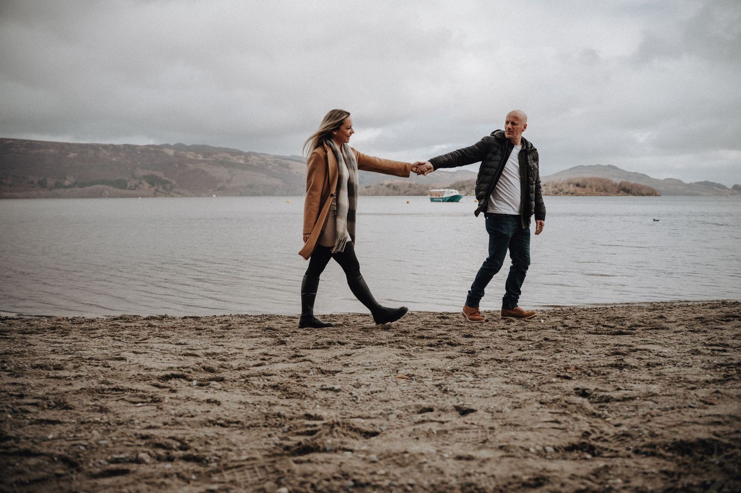 A couple holds hands while walking on a beach with mountains and moody skies in the background.