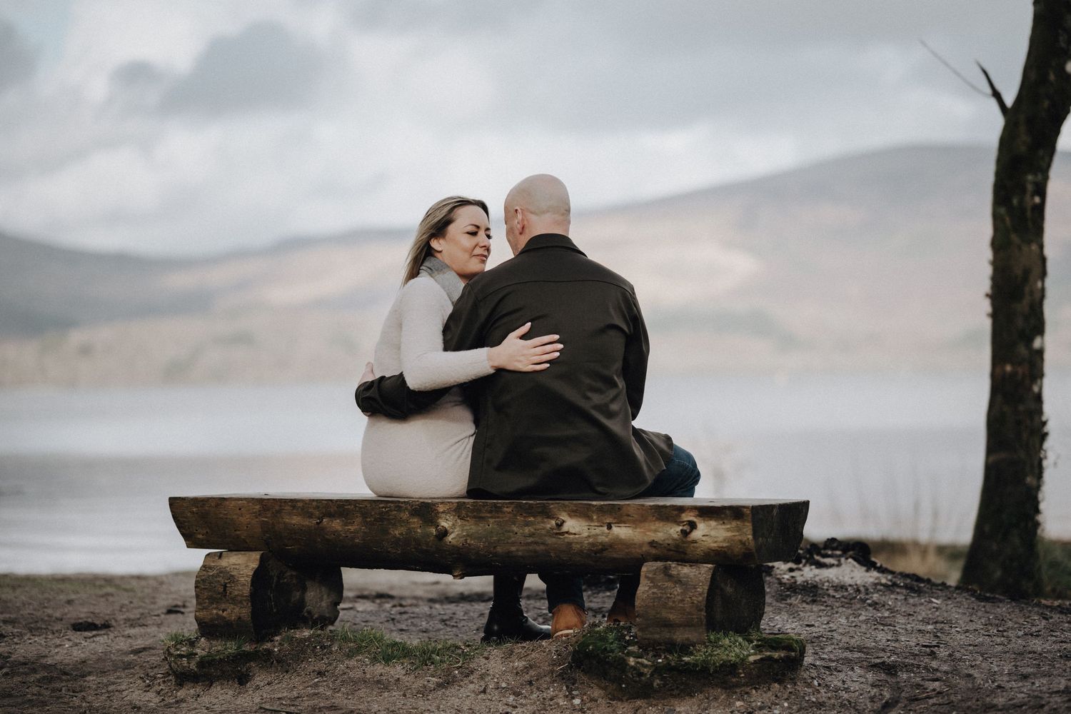 Intimate moment between couple embracing on bench with scenic mountain lake backdrop.