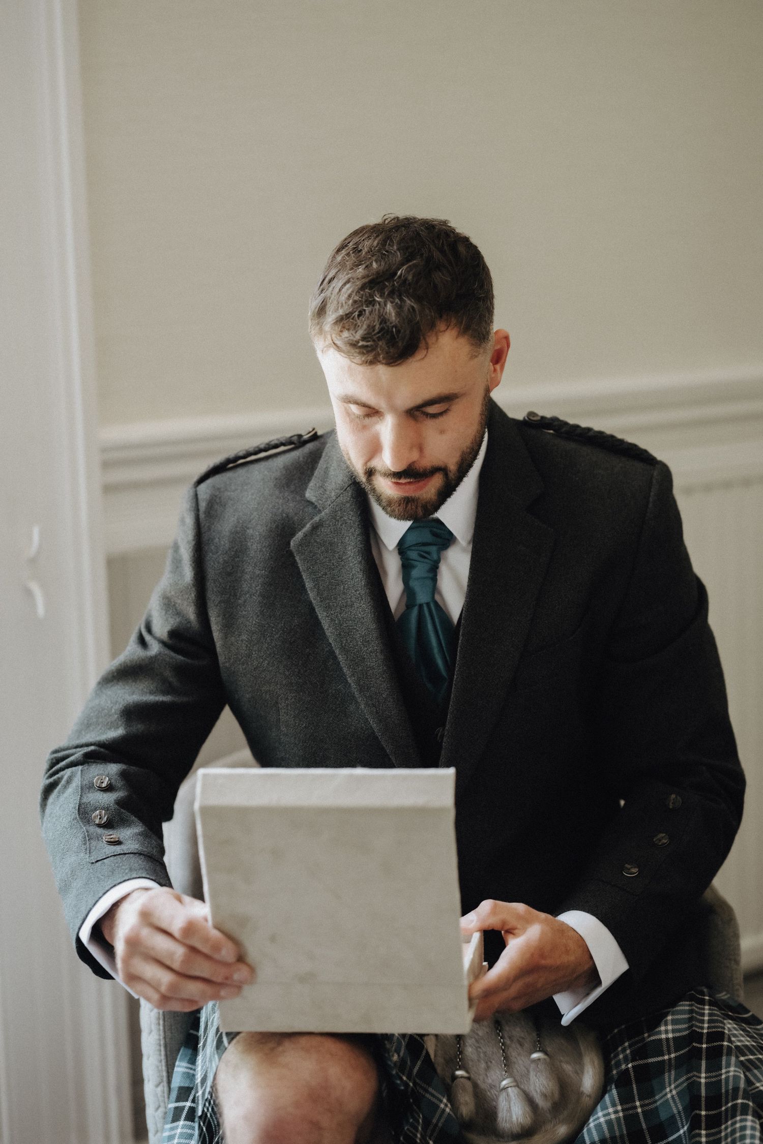 Person in formal Scottish attire sits reading from a white paper or card.