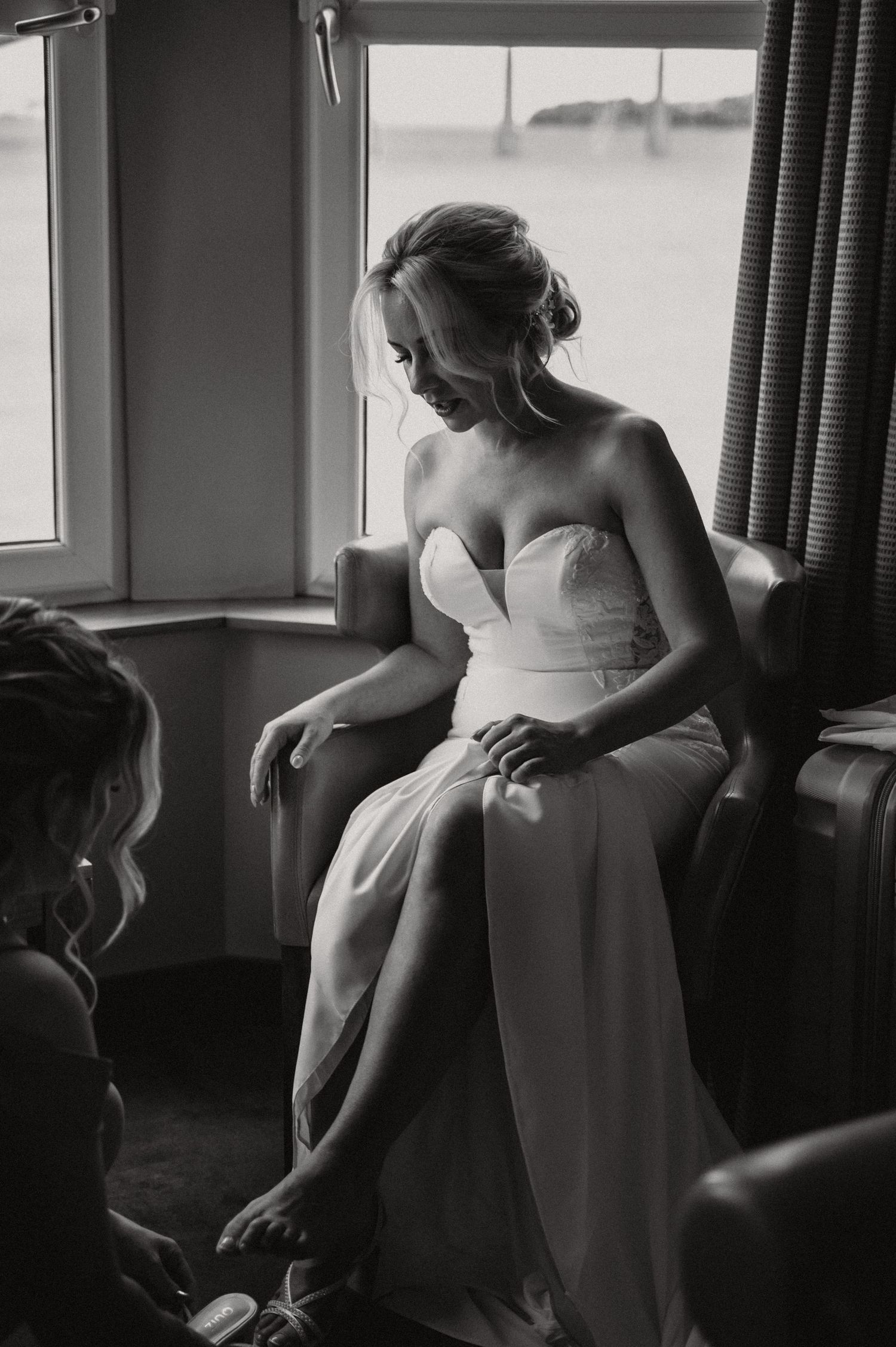 A bride in a white strapless gown sits by a window in a quiet moment before the ceremony.