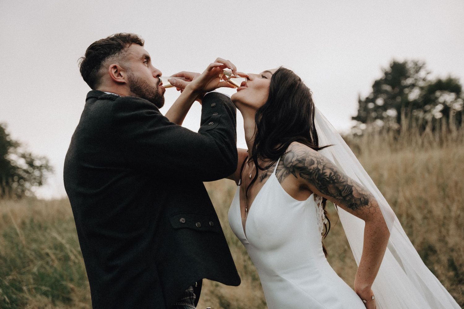 A playful couple shares champagne during their wedding photoshoot in an outdoor field.