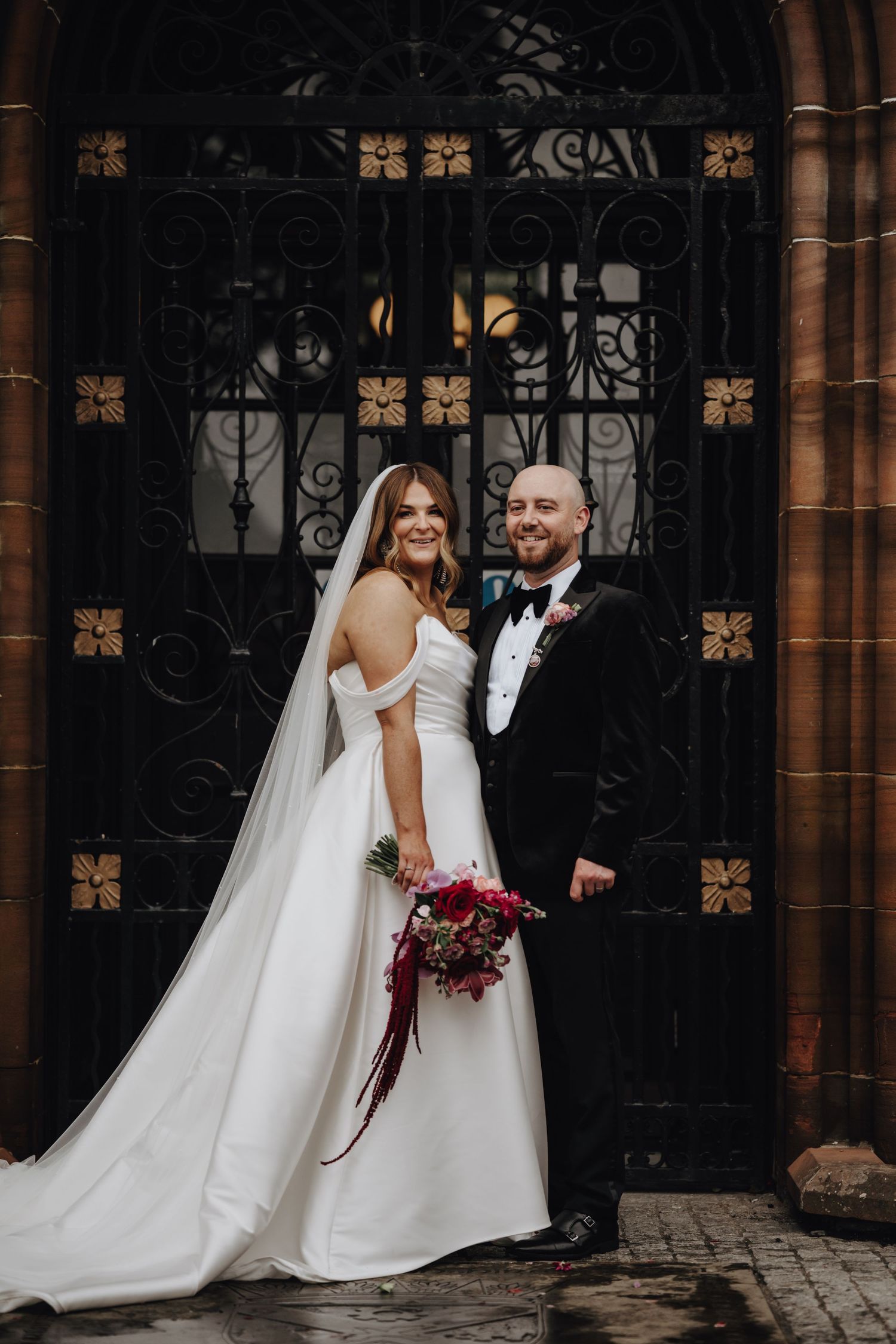 A couple poses in front of an ornate iron gate, with the bride holding a burgundy bouquet and wearing a flowing white gown.