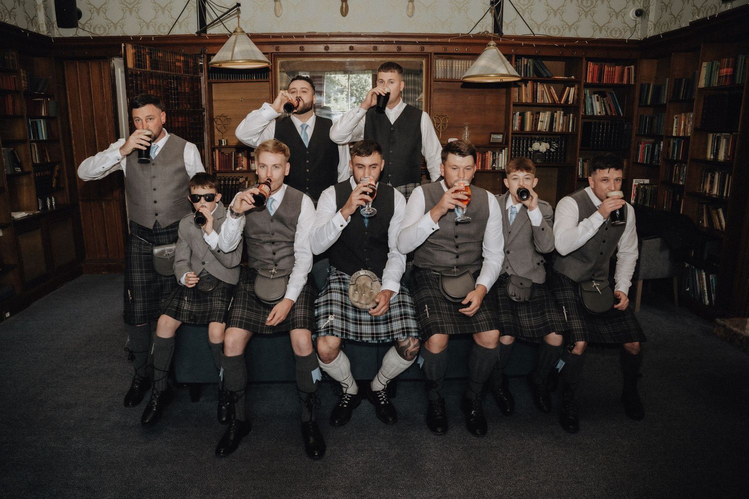 A group of people in kilts and waistcoats pose playfully in a library setting.