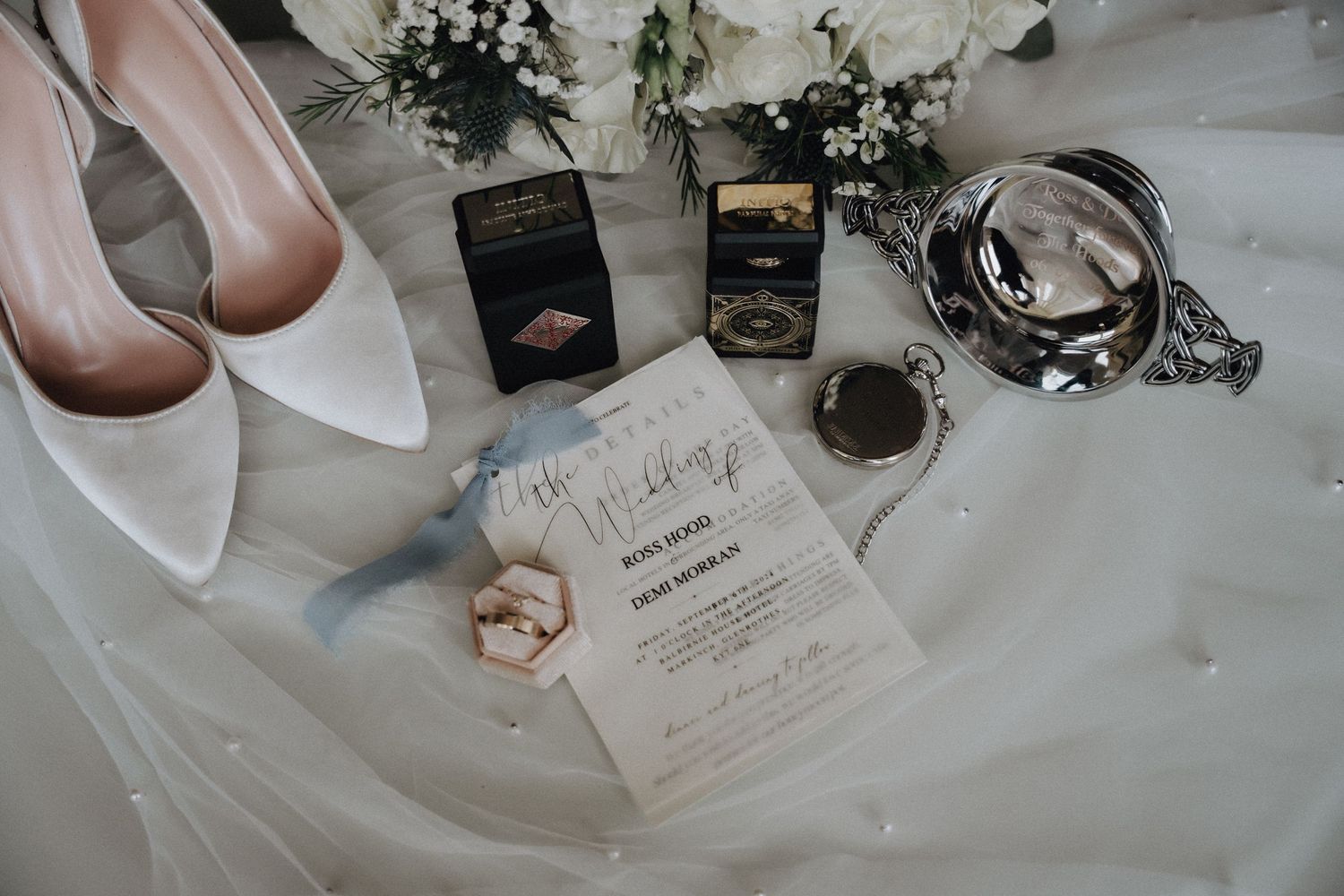 Wedding details flat lay with white heels, invitation, and accessories on delicate tulle fabric.