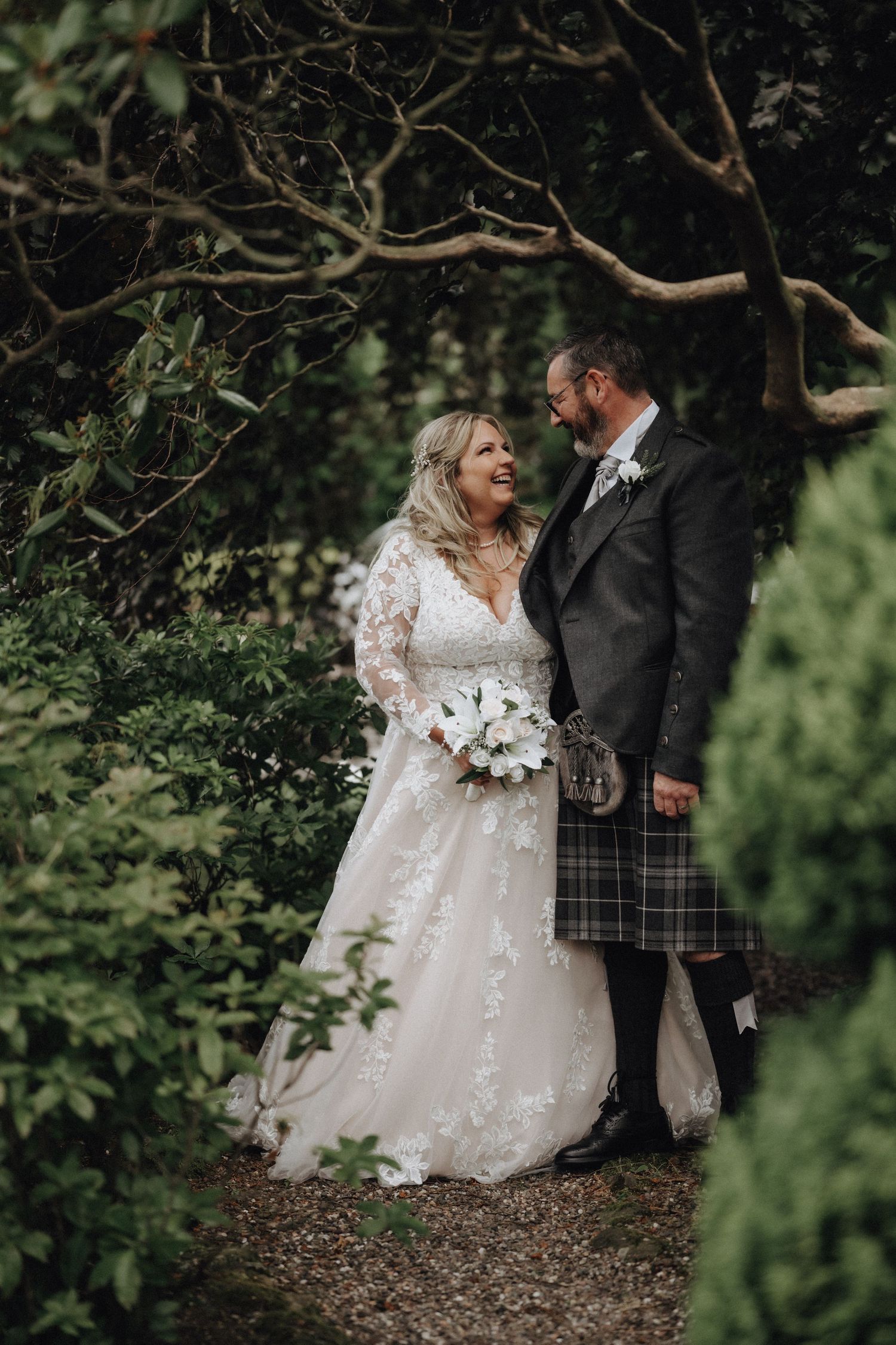 A romantic wedding couple poses in a garden setting with the groom wearing a traditional Scottish kilt.