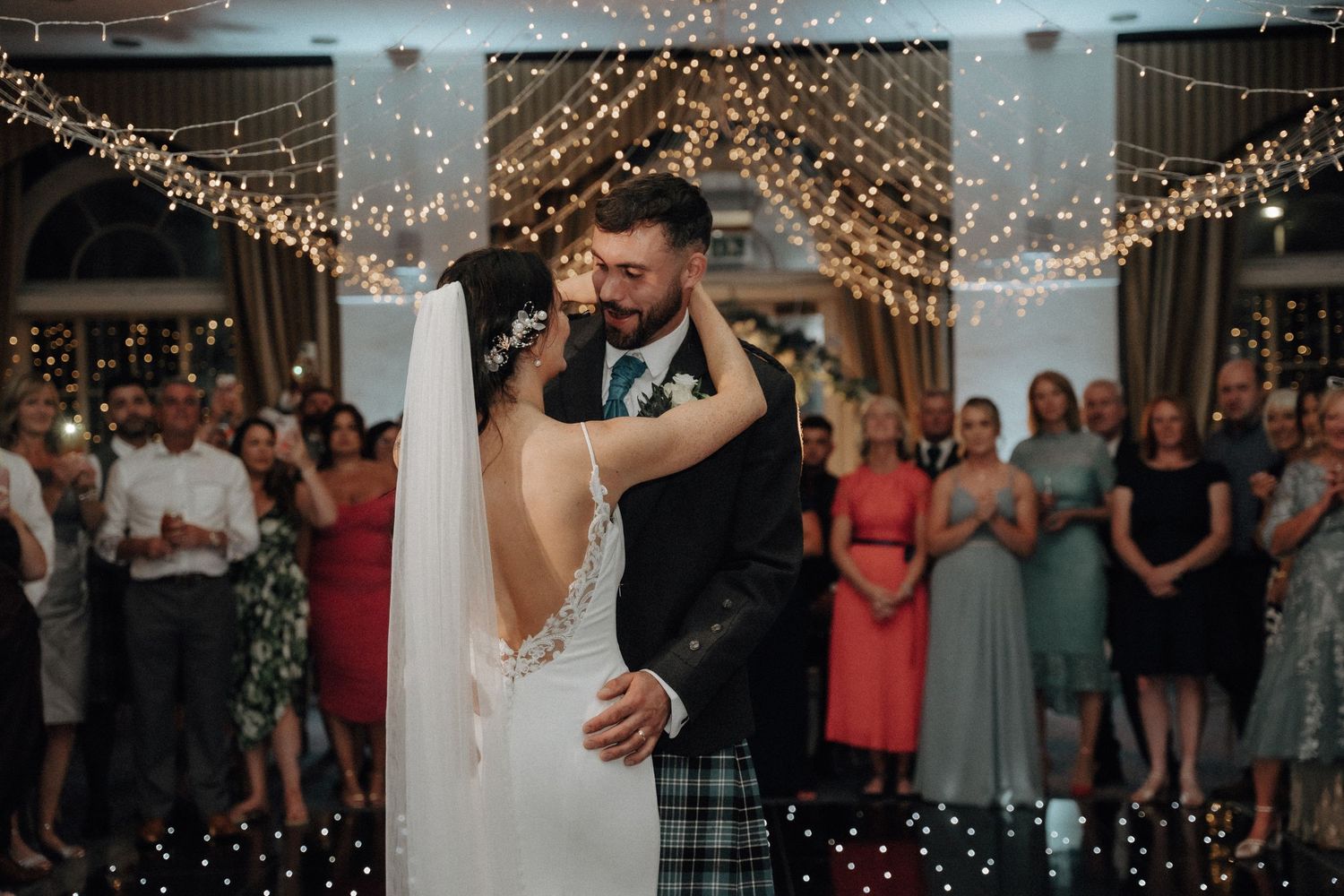 First dance of newlyweds under romantic twinkle lights with guests watching.