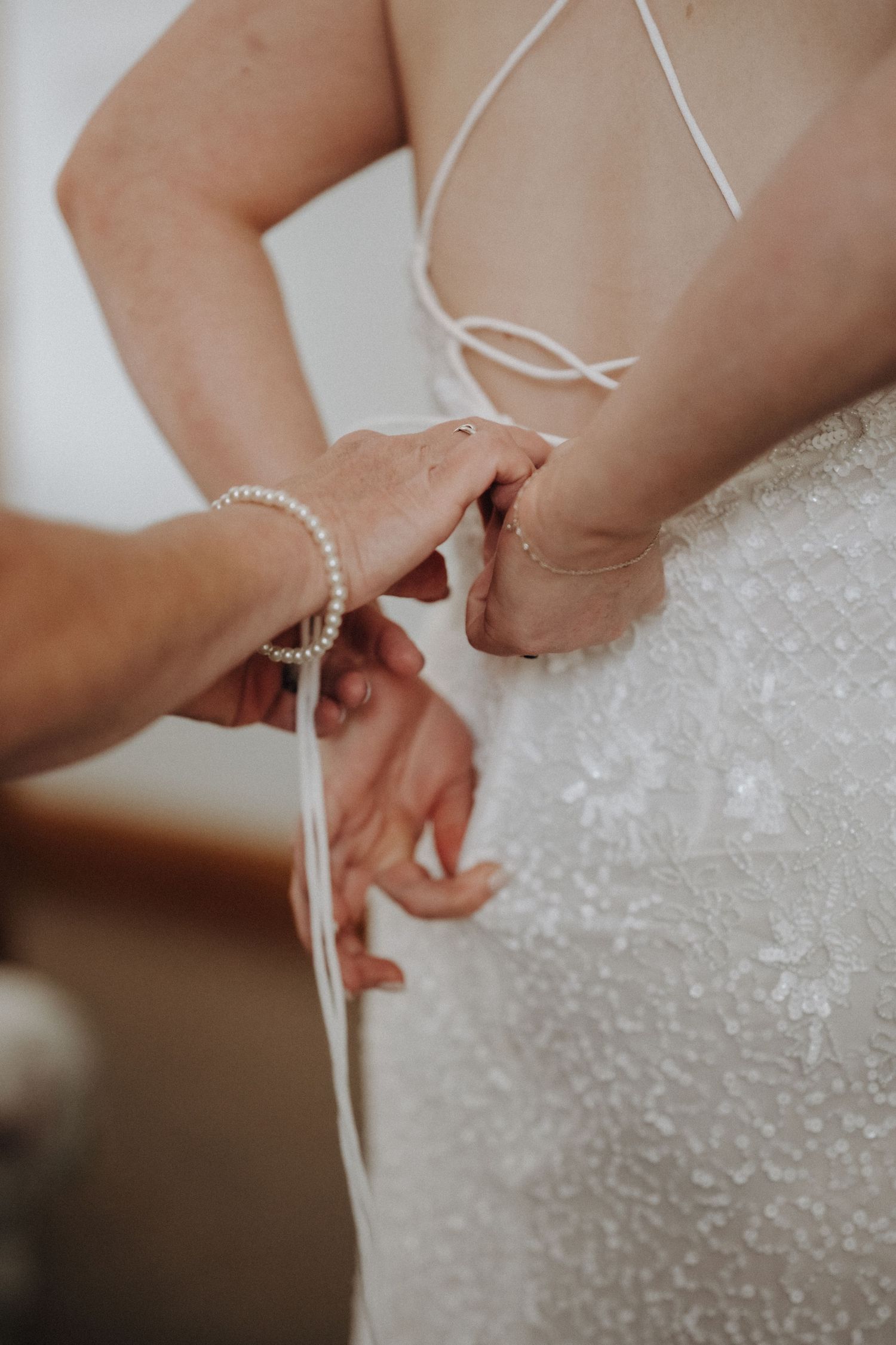 Close-up of hands helping bride fasten white dress with pearl bracelet detail.