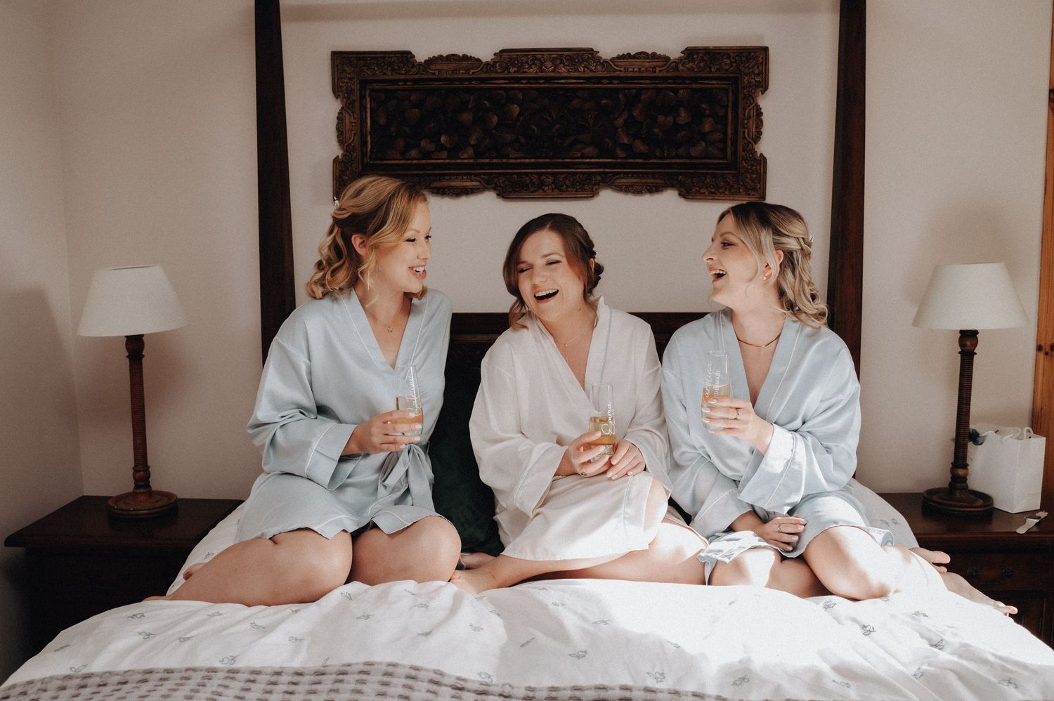 Three friends in matching robes sitting on bed laughing together during wedding preparations.