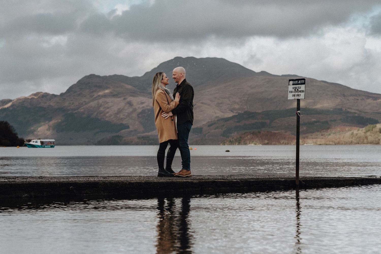 A couple stands together on a pier overlooking a misty Scottish loch with mountains in the distance.