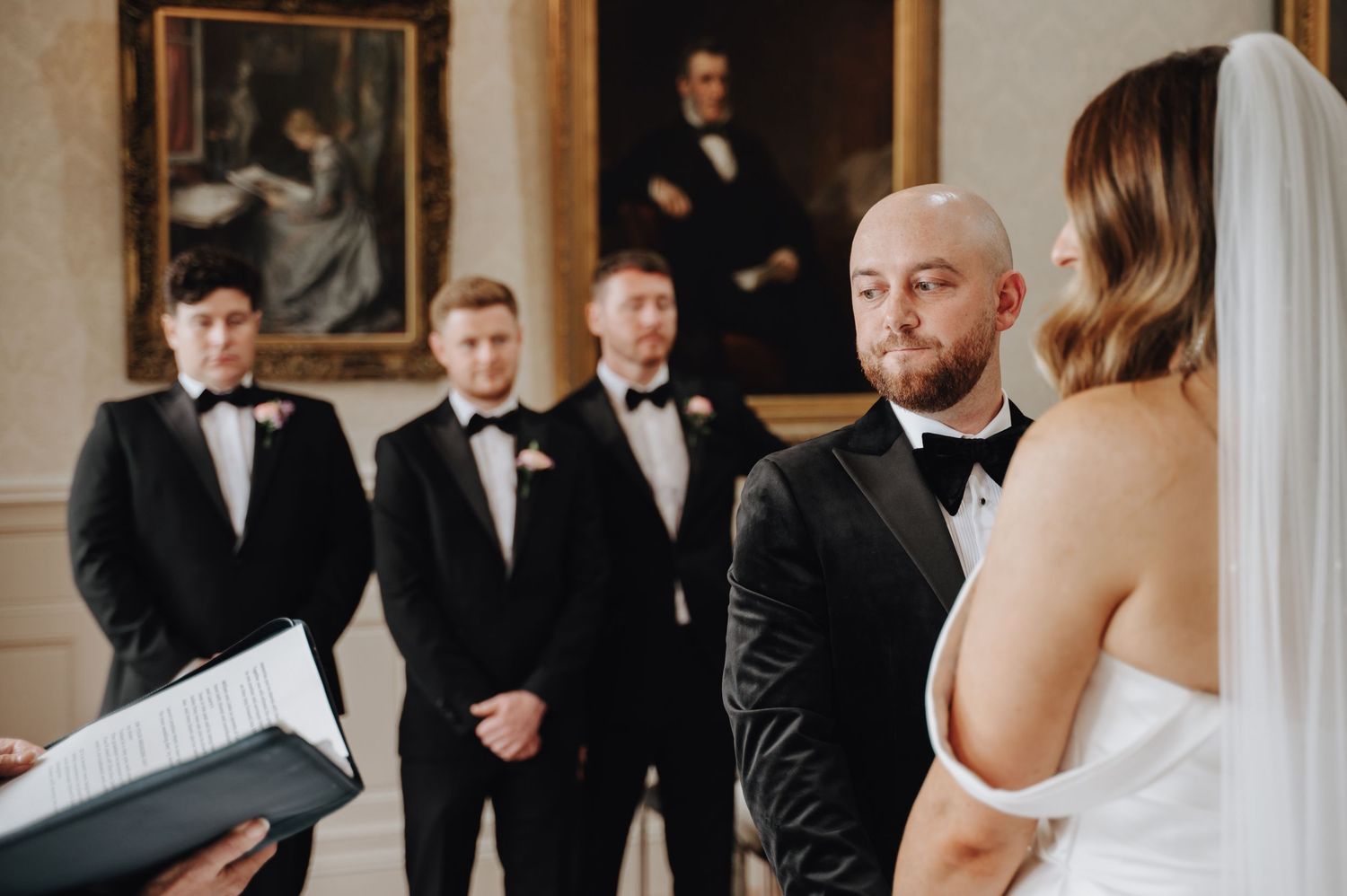 Wedding party awaits in a grand room with ornate portrait art and classic architectural details as couple shares a moment.