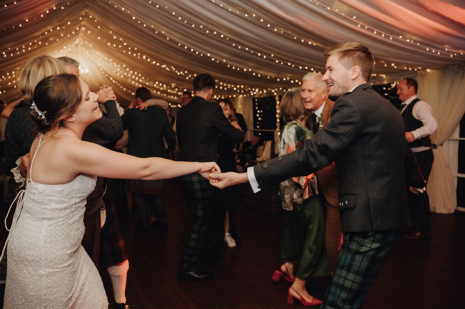 Wedding guests dancing in a tent decorated with twinkling lights and elegant draping.