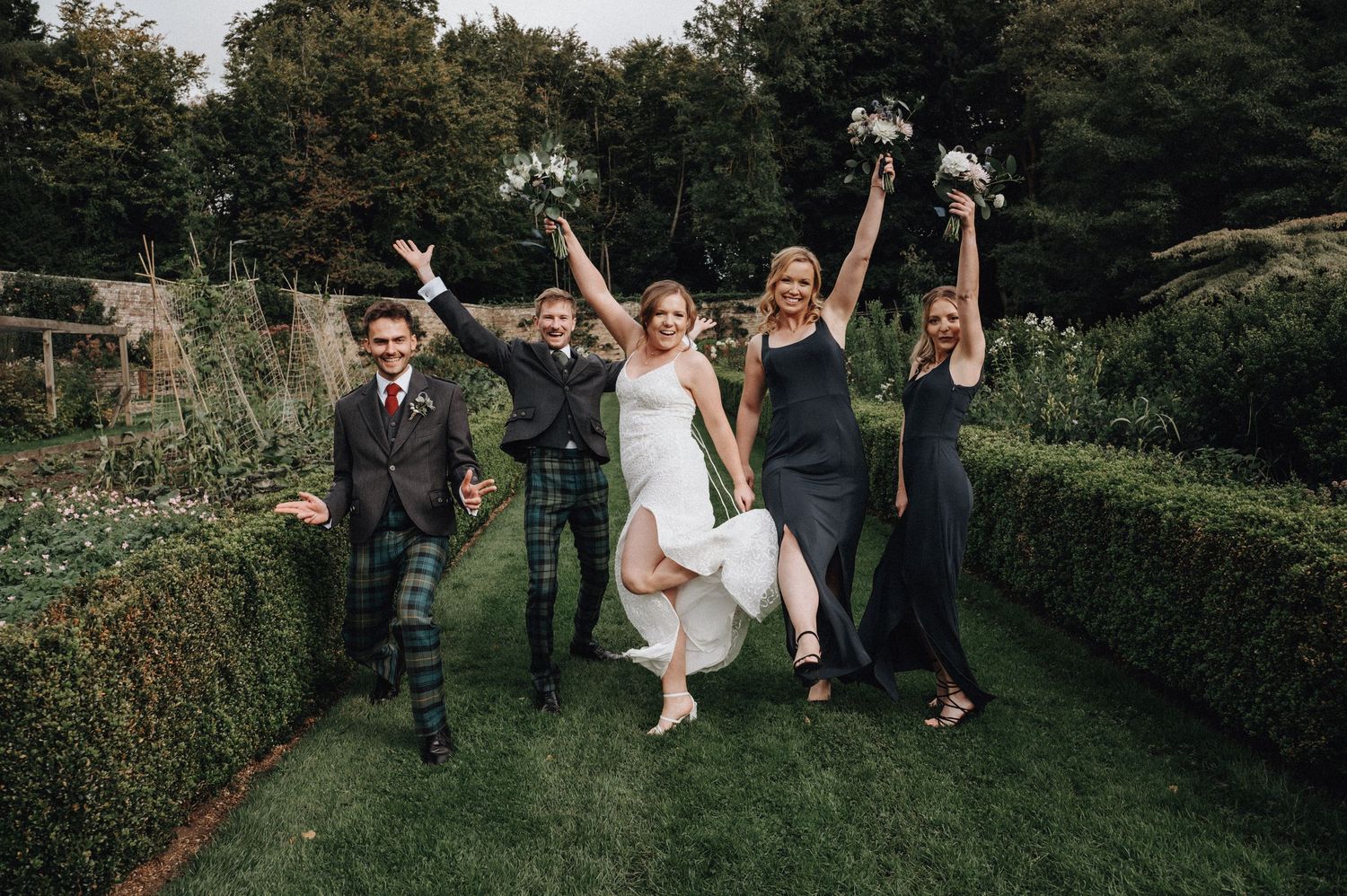 Wedding party celebrates joyfully in a formal garden setting surrounded by hedges.