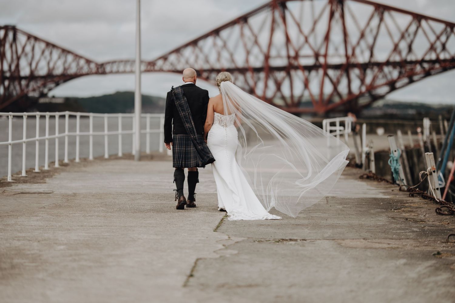 Bride's veil flows in wind as couple walks along pier with bridge in background.