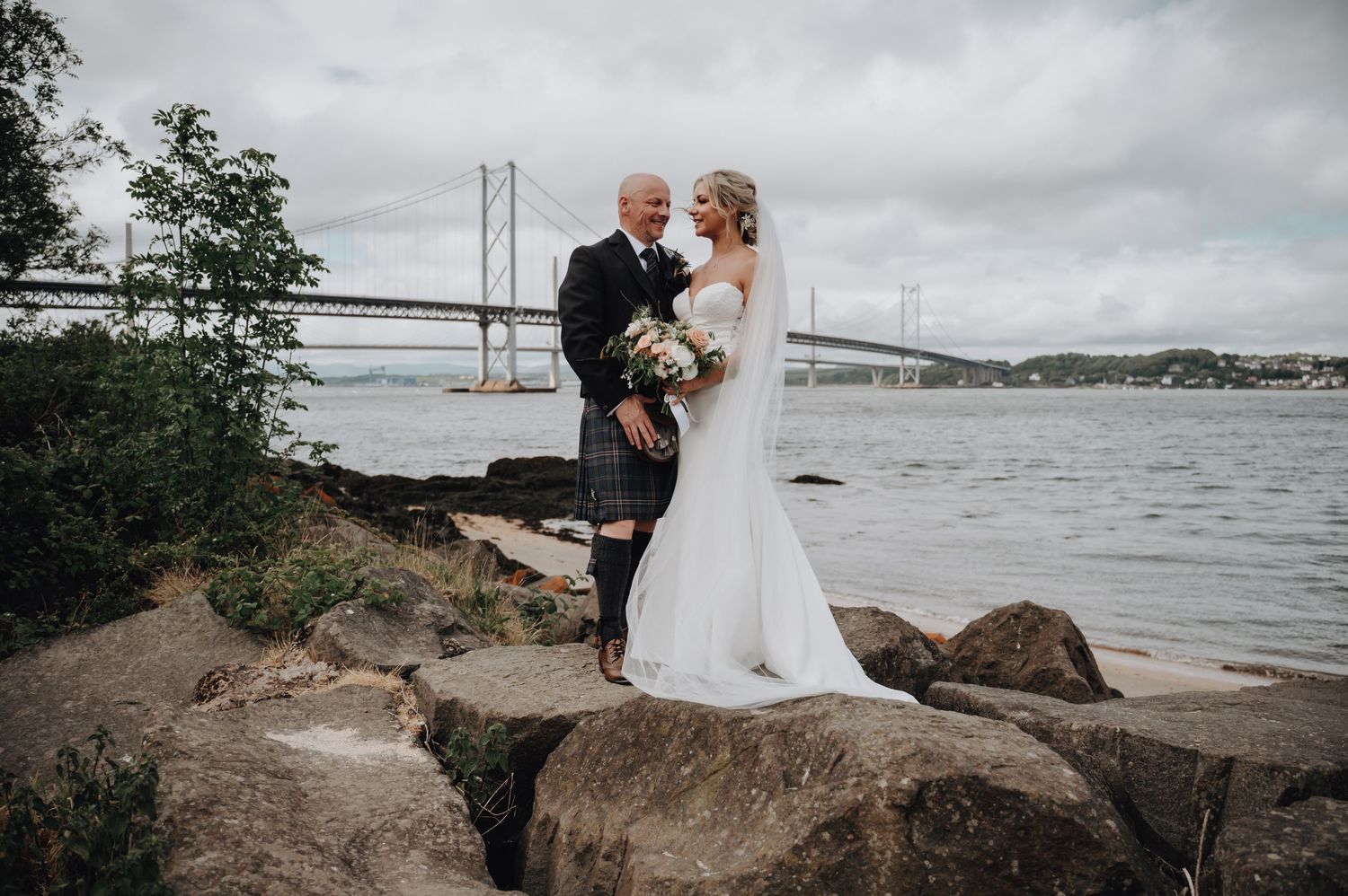 Wedding couple stands on rocky shoreline with bridges visible across water on cloudy day.