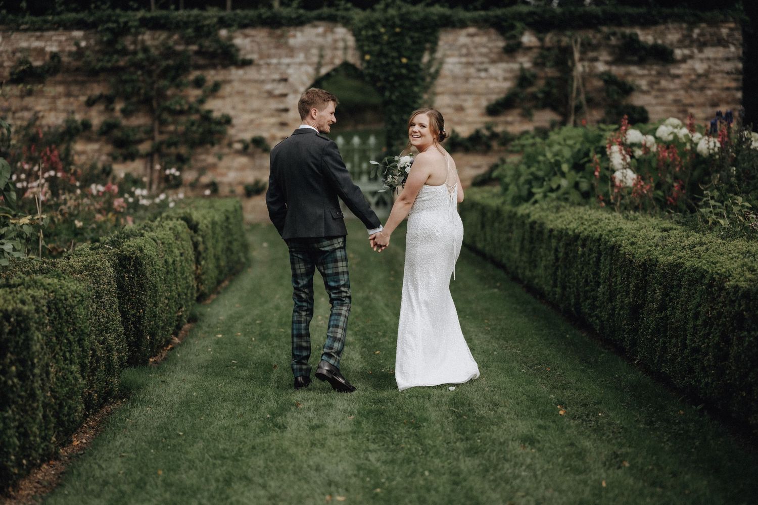 Couple poses together in formal wear on a manicured garden path surrounded by hedges.
