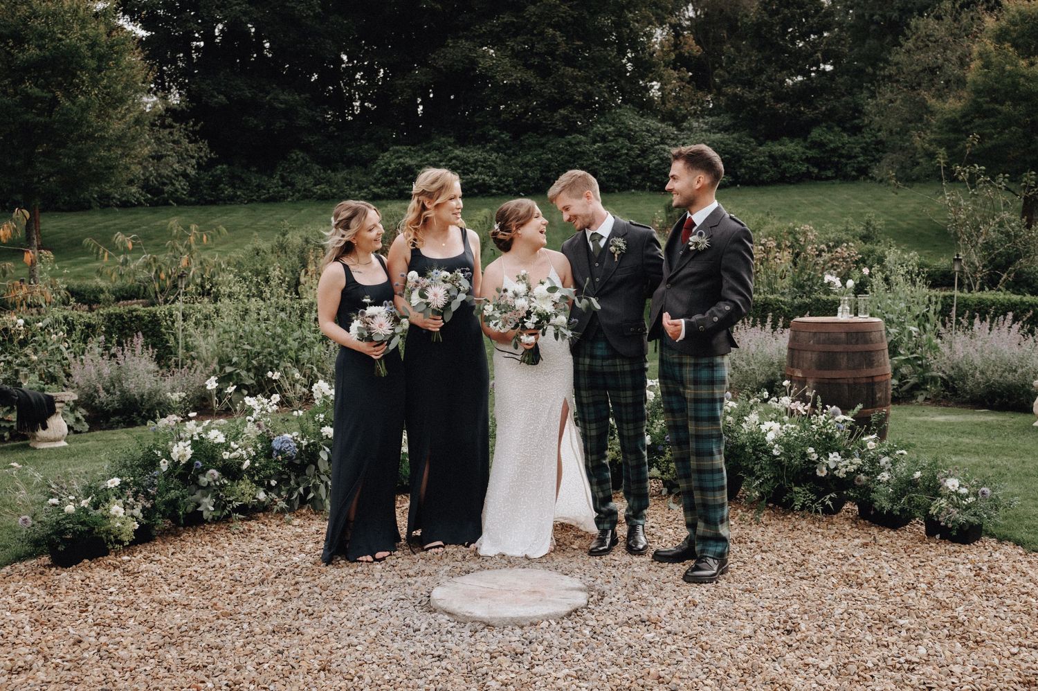 Wedding party poses together in a garden setting with traditional Scottish attire and bridal bouquets.