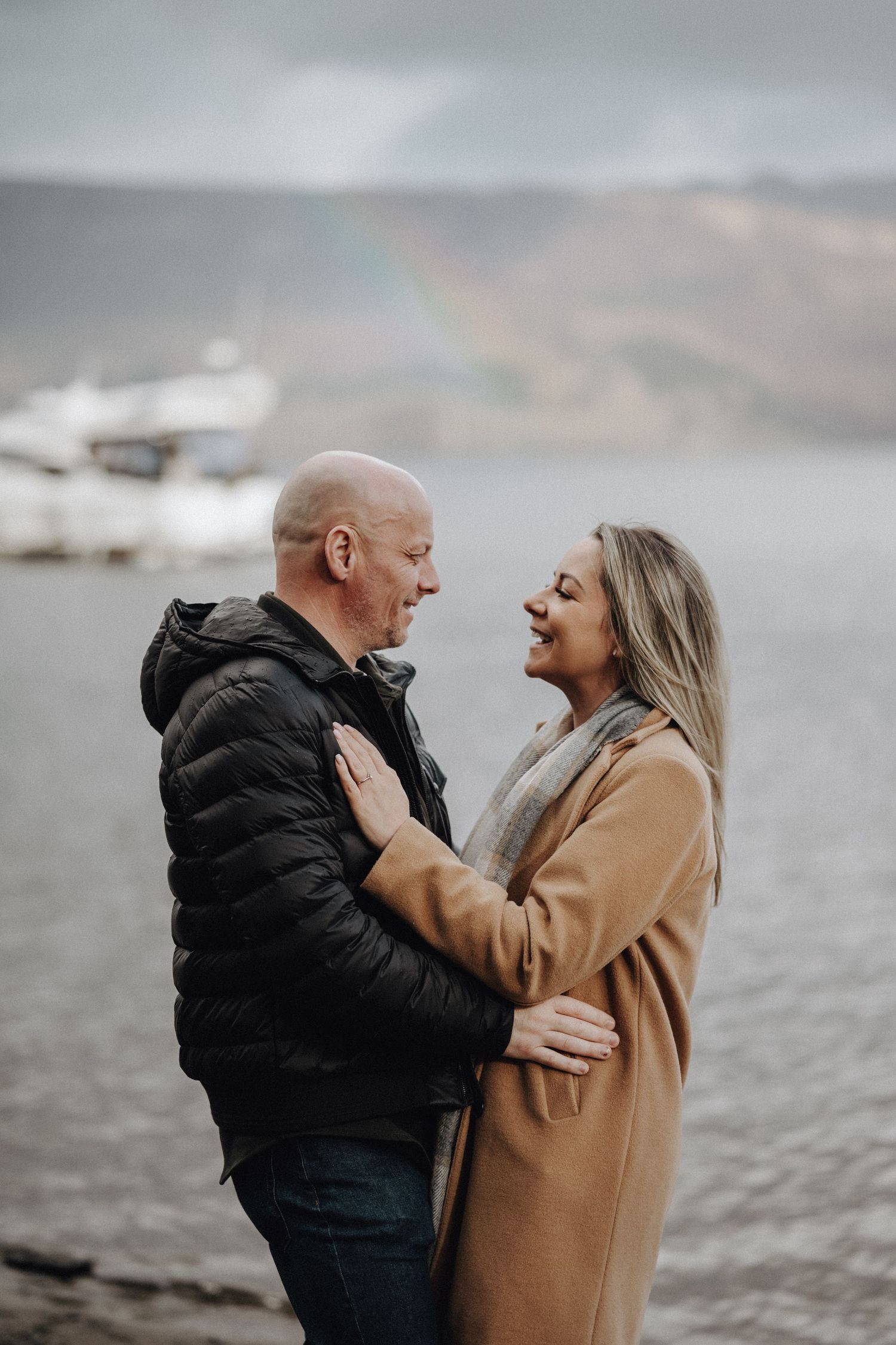 A couple laughs together while standing on a beach with a rainbow visible in the distance.
