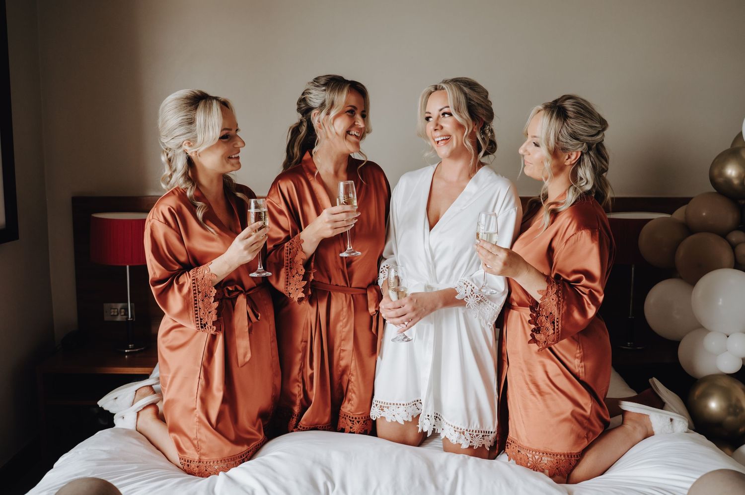 Wedding party in contrasting silk robes laugh together on hotel bed during preparations.
