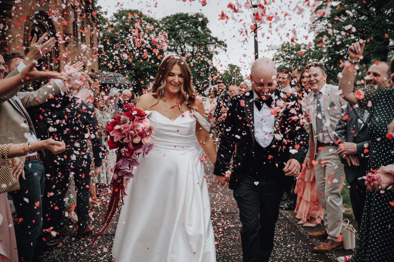 Smiling wedding party is showered with pink and white confetti petals during outdoor celebration festivities.