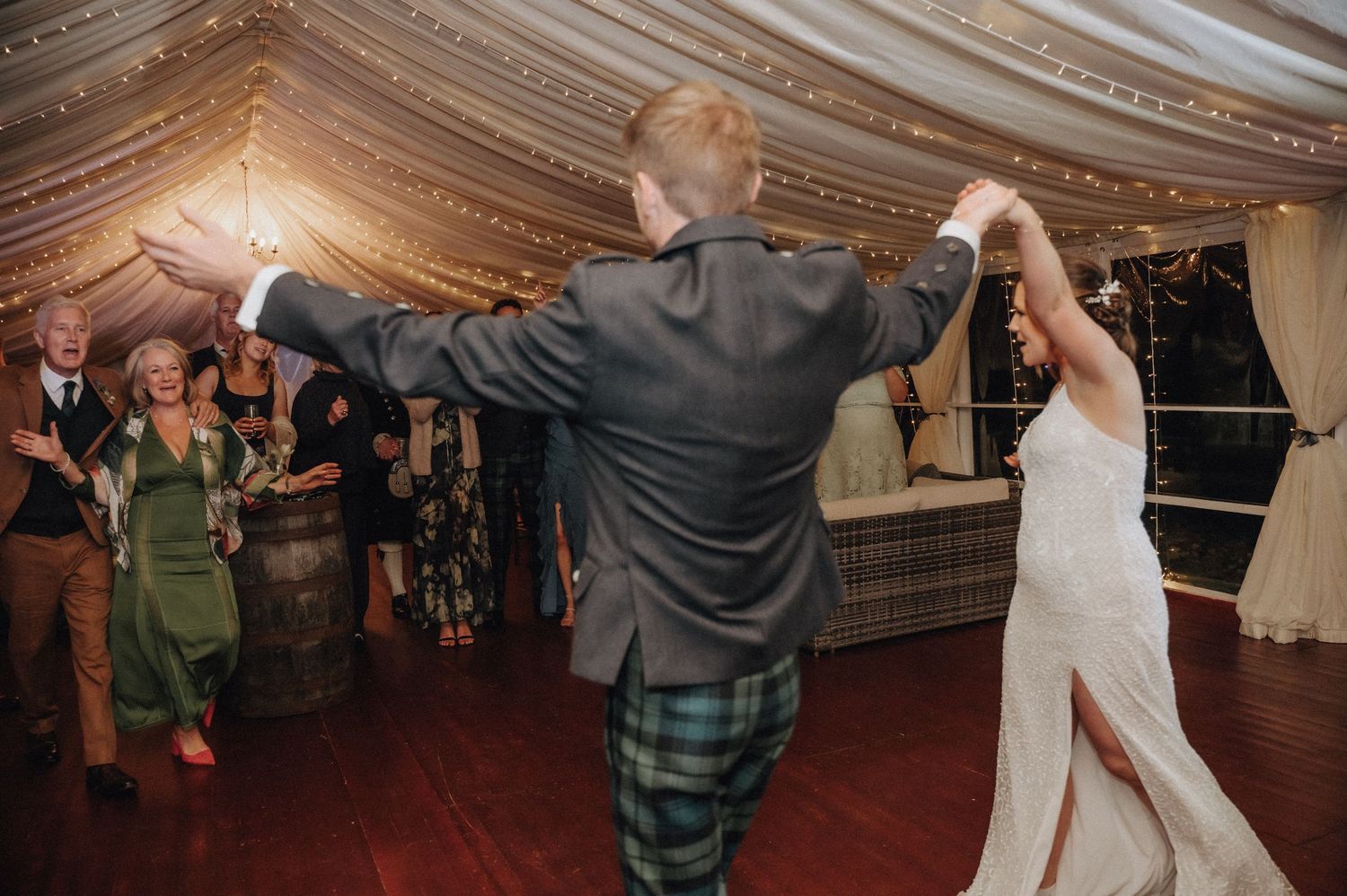 Wedding celebration with dancing guests in traditional Scottish attire under fairy-lit tent ceiling.