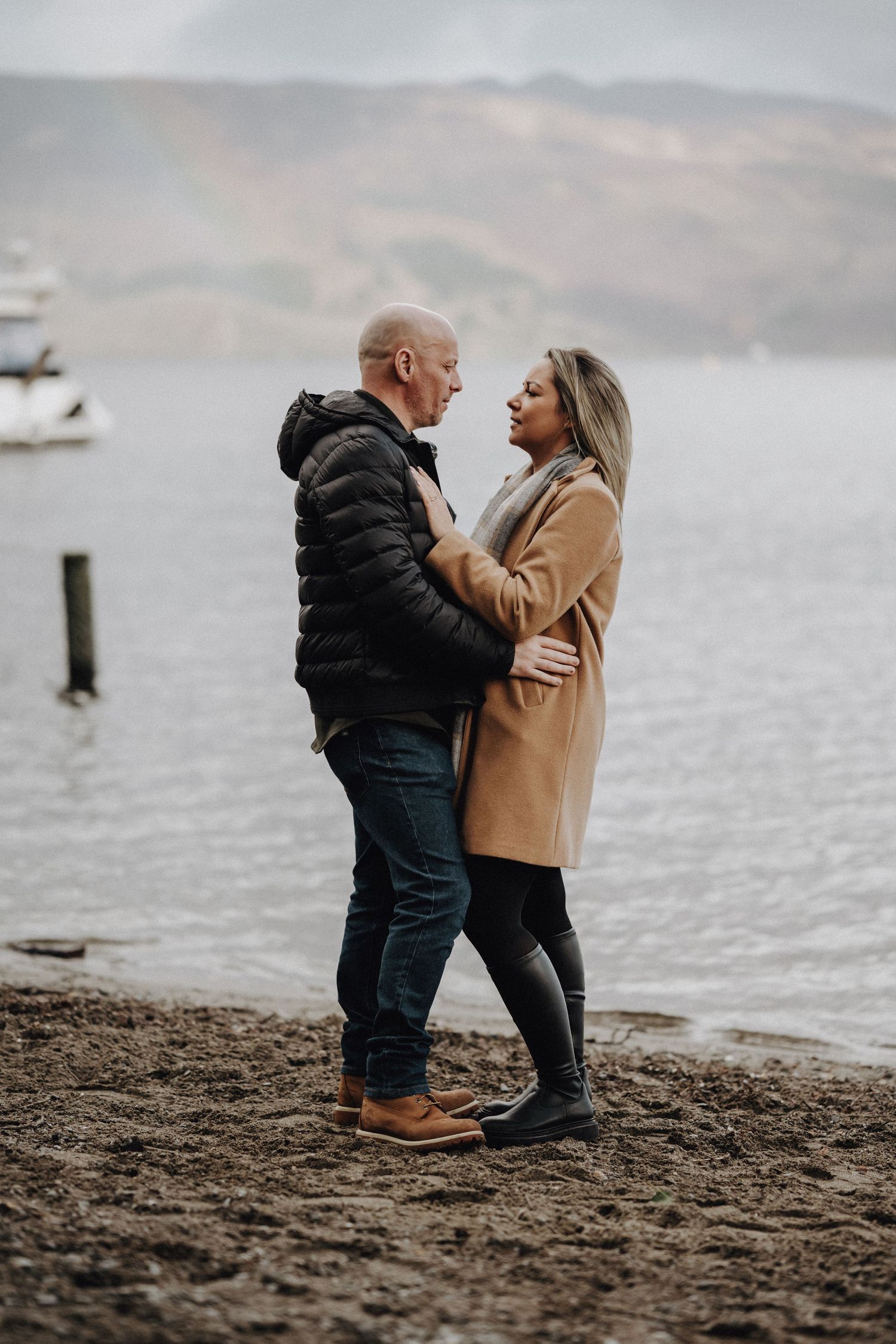 A couple shares an intimate moment on a beach with boats and mountains in the background.