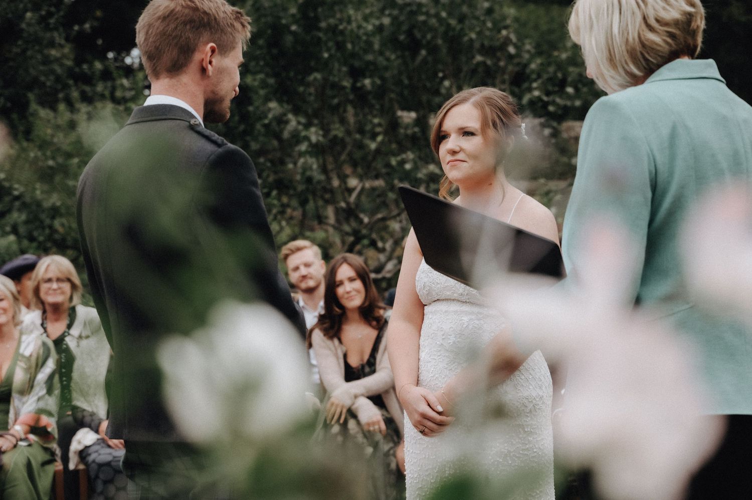 Intimate outdoor wedding ceremony with guests seated on lawn near greenhouse.
