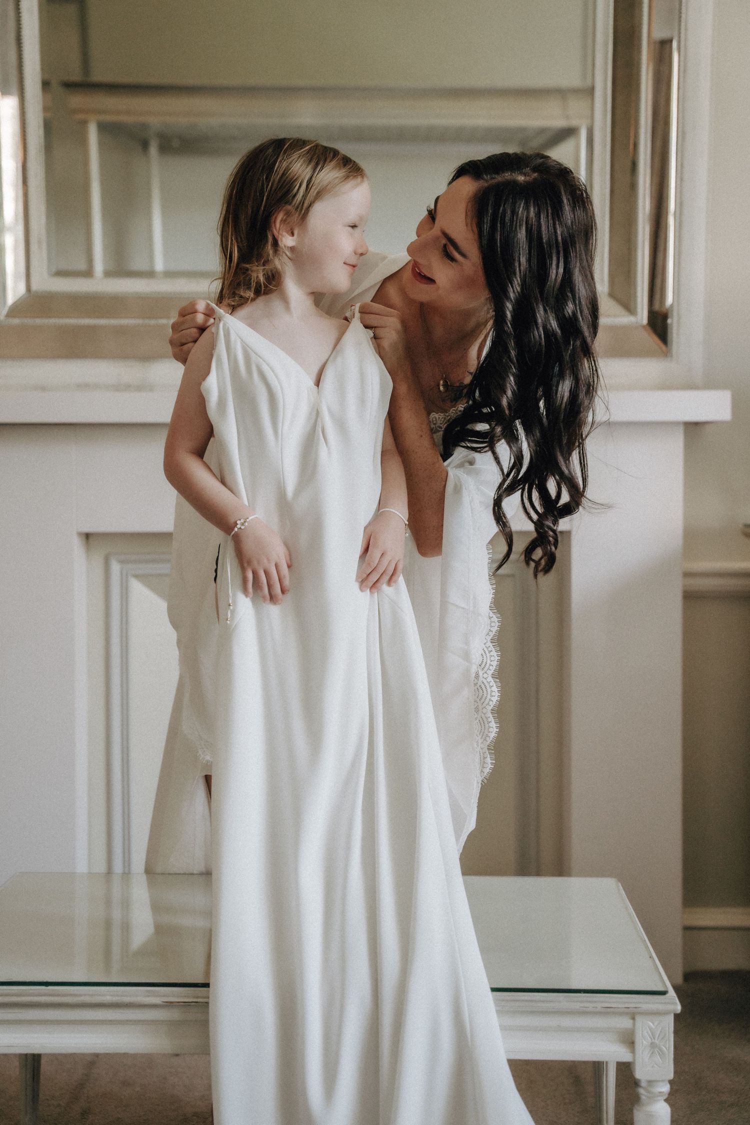 A tender moment shared between a woman in white and a young child in a white dress standing on a table.