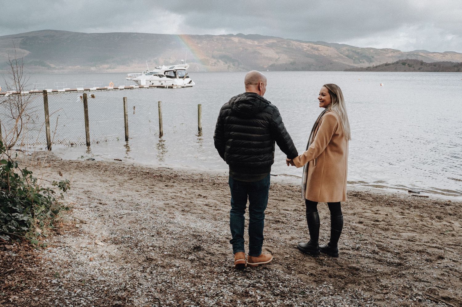 A couple holding hands watches a plane fly over a calm lake surrounded by mountains.