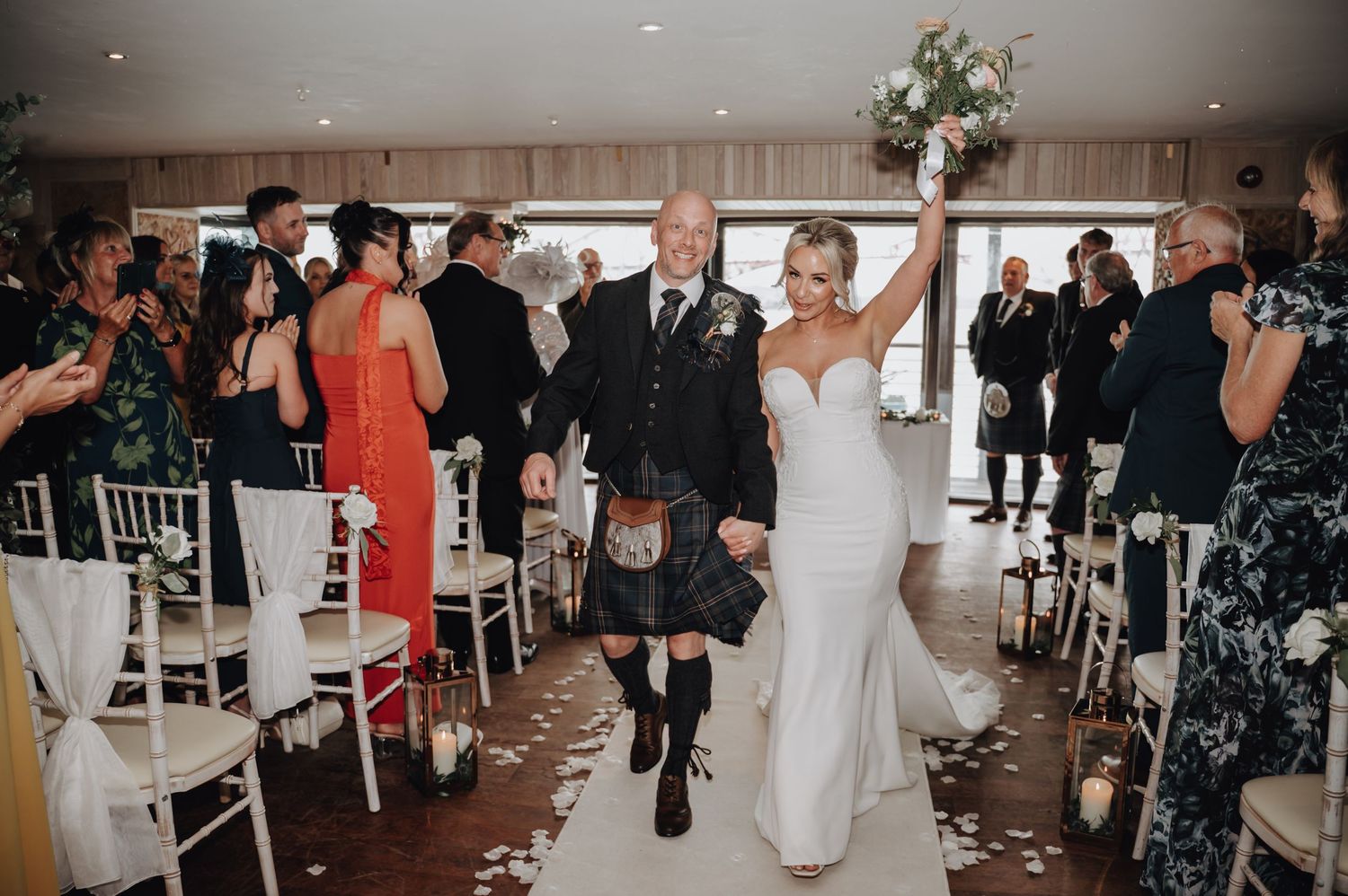 Newlyweds joyfully exit down the aisle at a wedding ceremony with guests celebrating in white chairs.