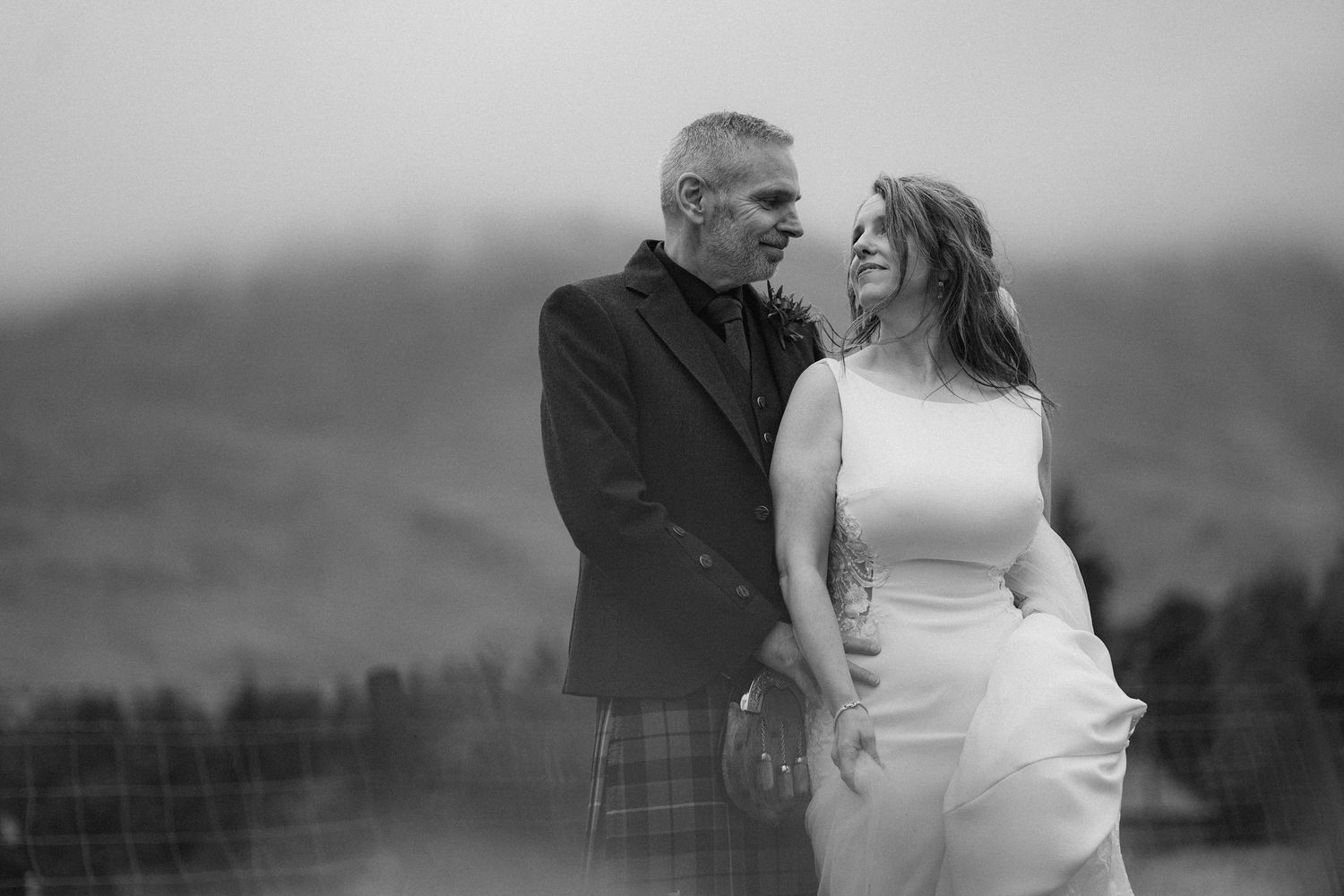 A couple in wedding attire shares a joyful moment against a misty Scottish highland backdrop.