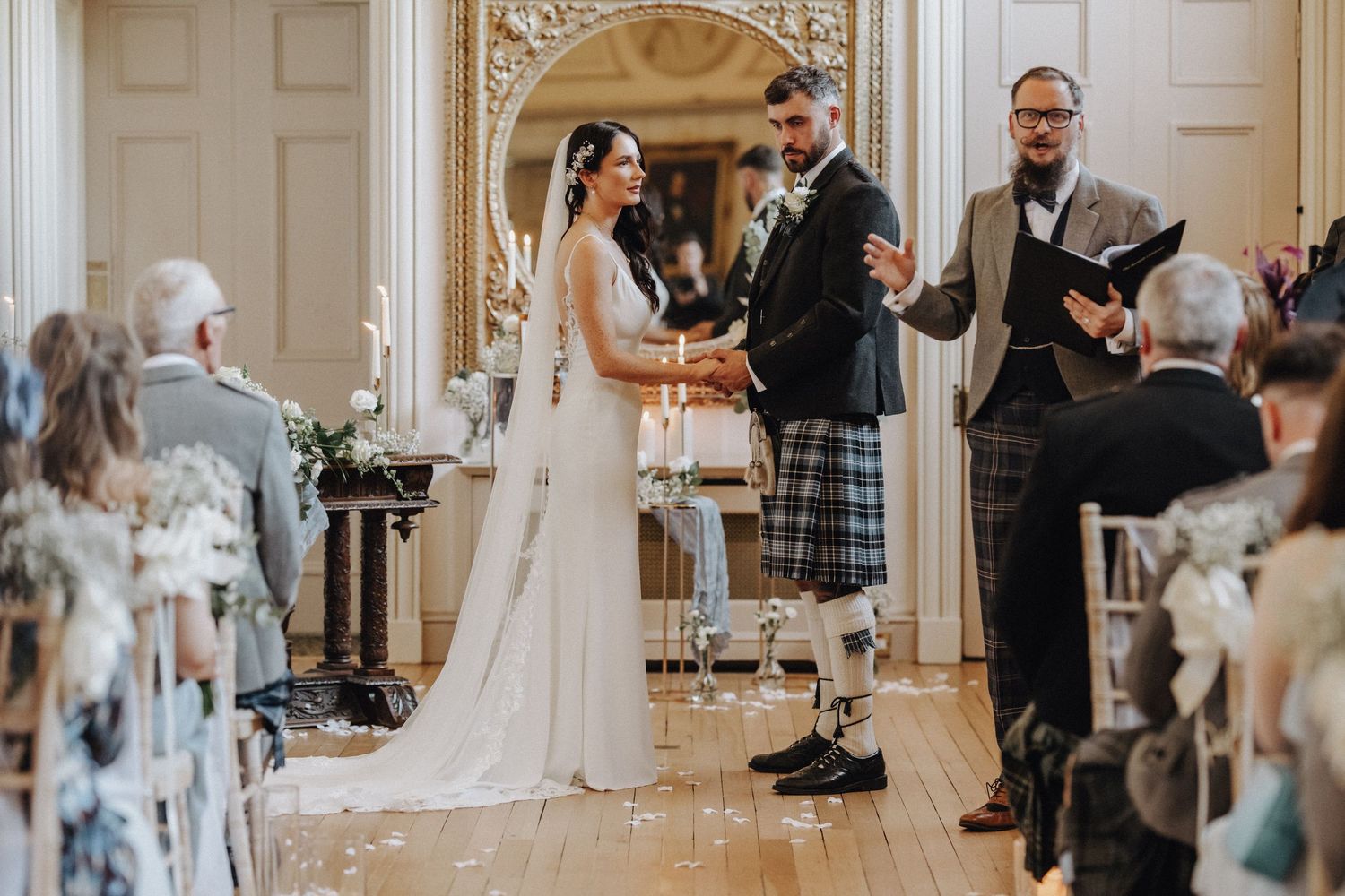 Wedding ceremony in progress with couple and officiant standing before seated guests in an elegant room.