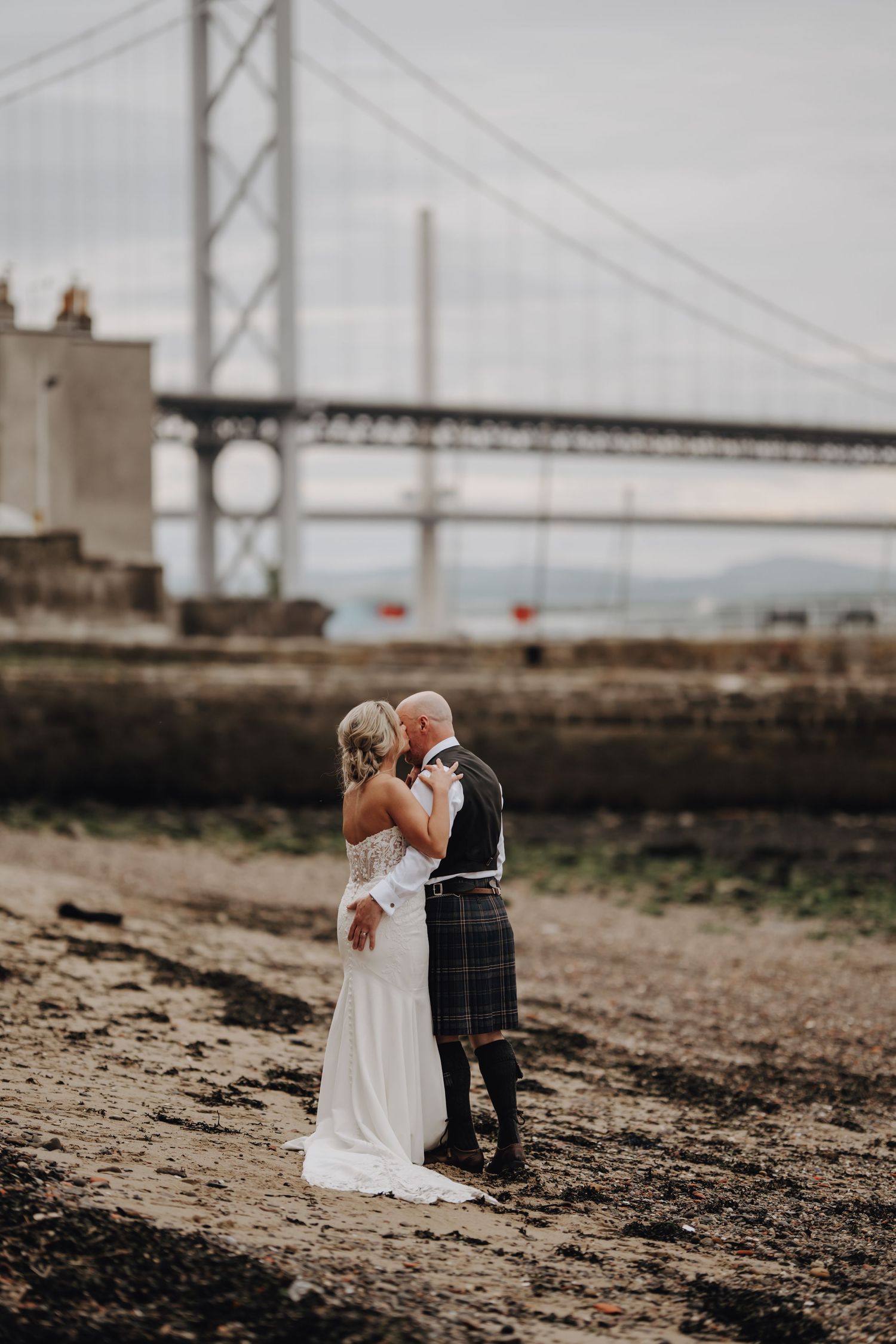 Romantic wedding photo taken by a bridge with couple on rocky beach.