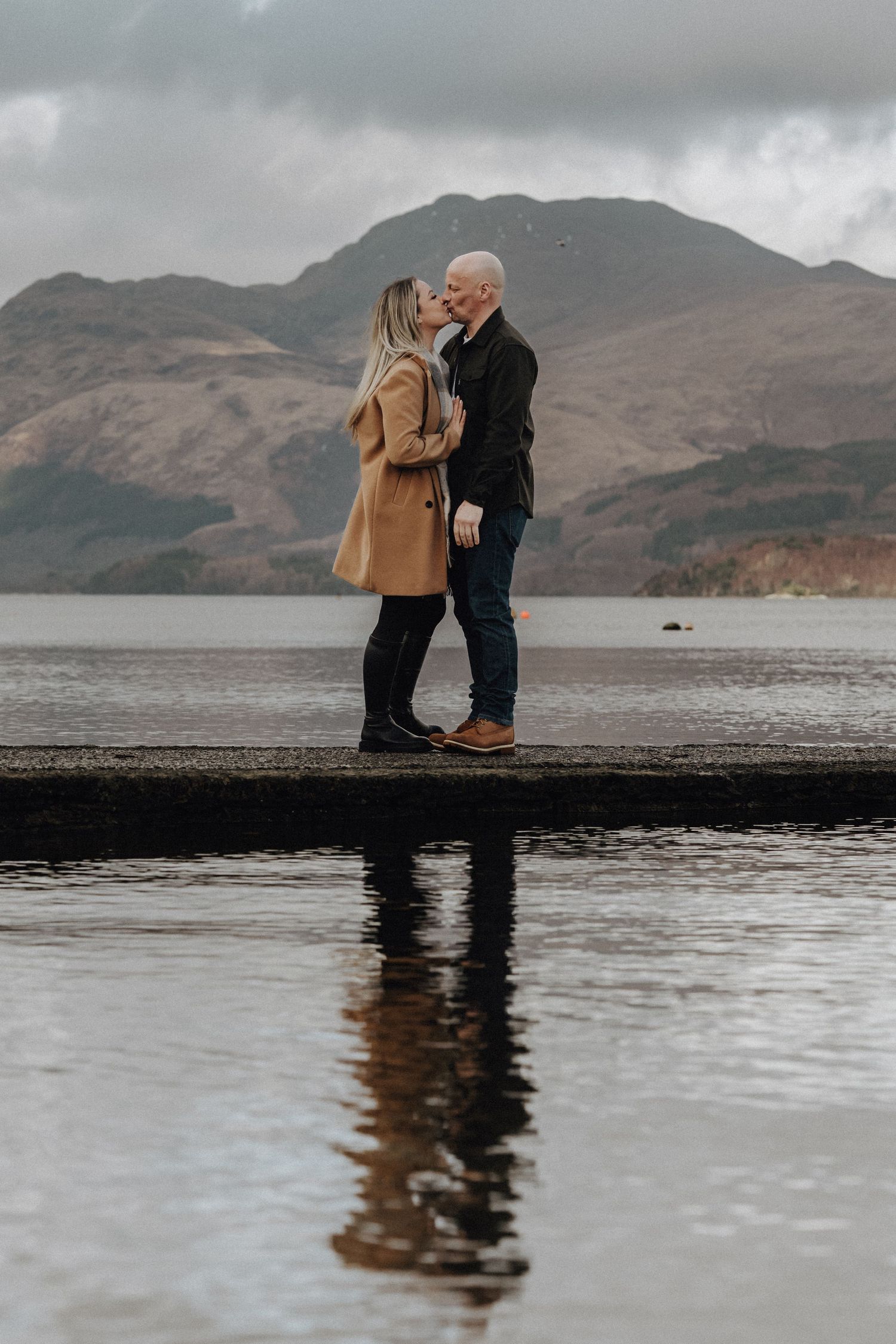 A couple stands close together on a dock with misty mountains and reflective water behind them.
