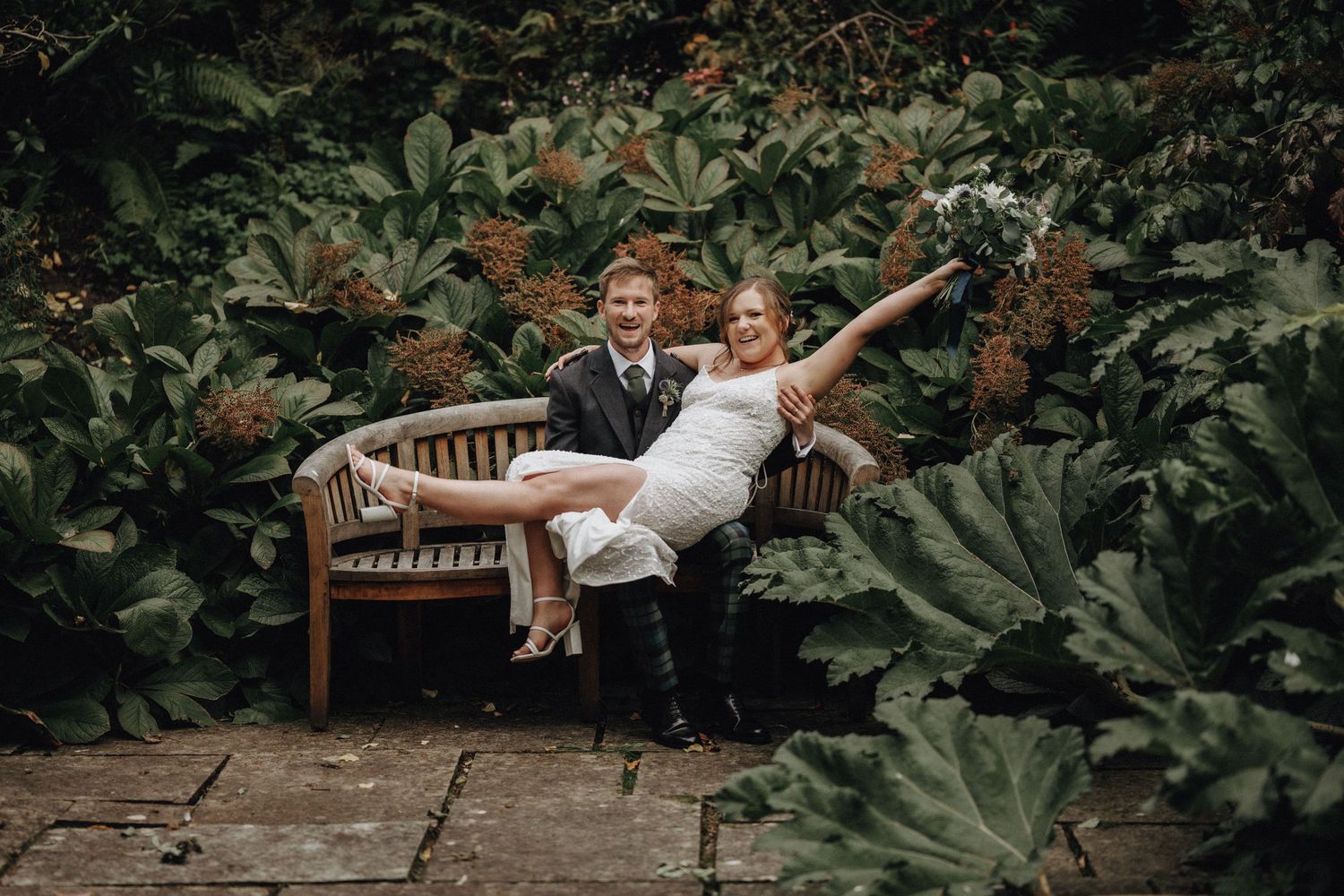Playful newlyweds pose together on wooden garden bench surrounded by foliage.
