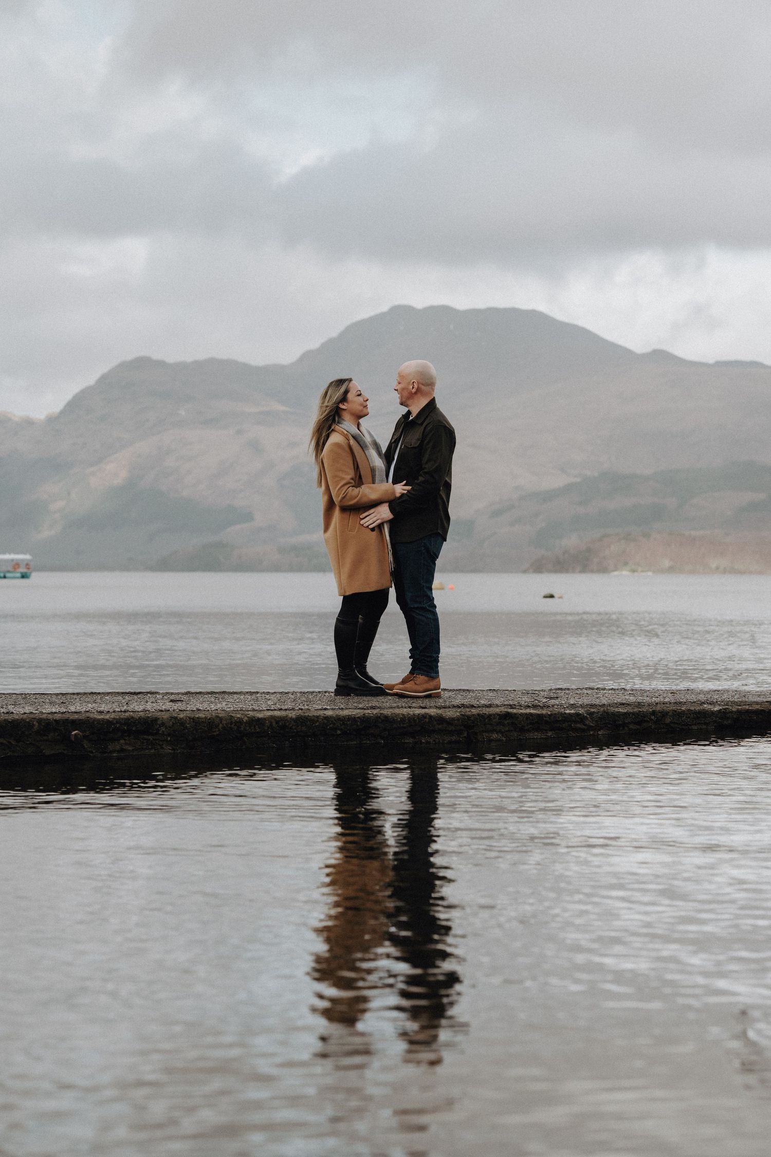 Romantic couple standing close on a pier with mountains and cloudy skies in the background.