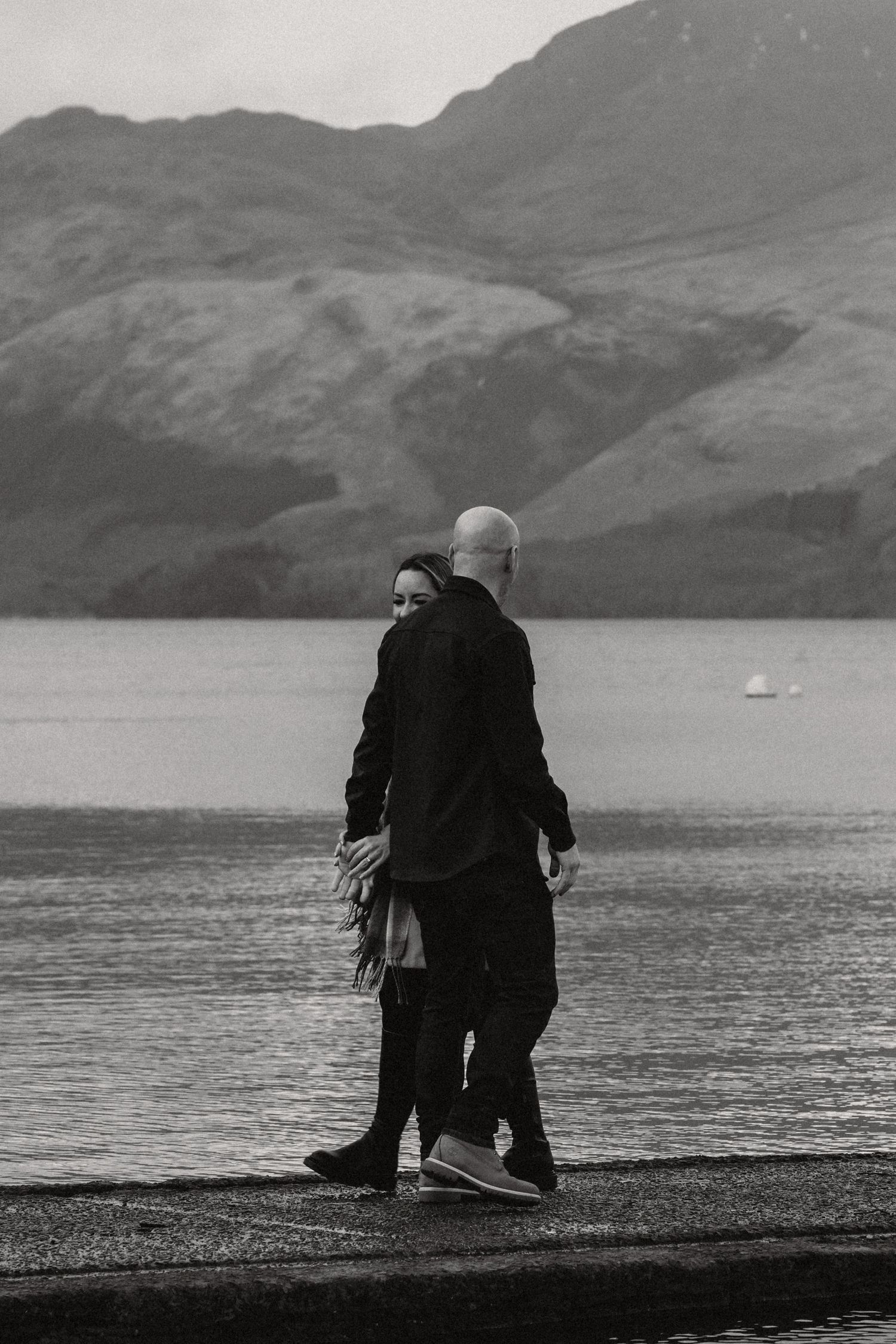 A black and white photo of a couple standing by a serene lake with mountains in the background.