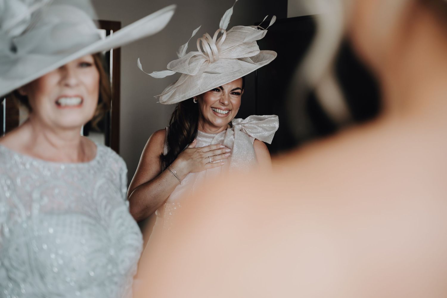 Wedding guests wearing elegant hats and dresses smile during a formal celebration.
