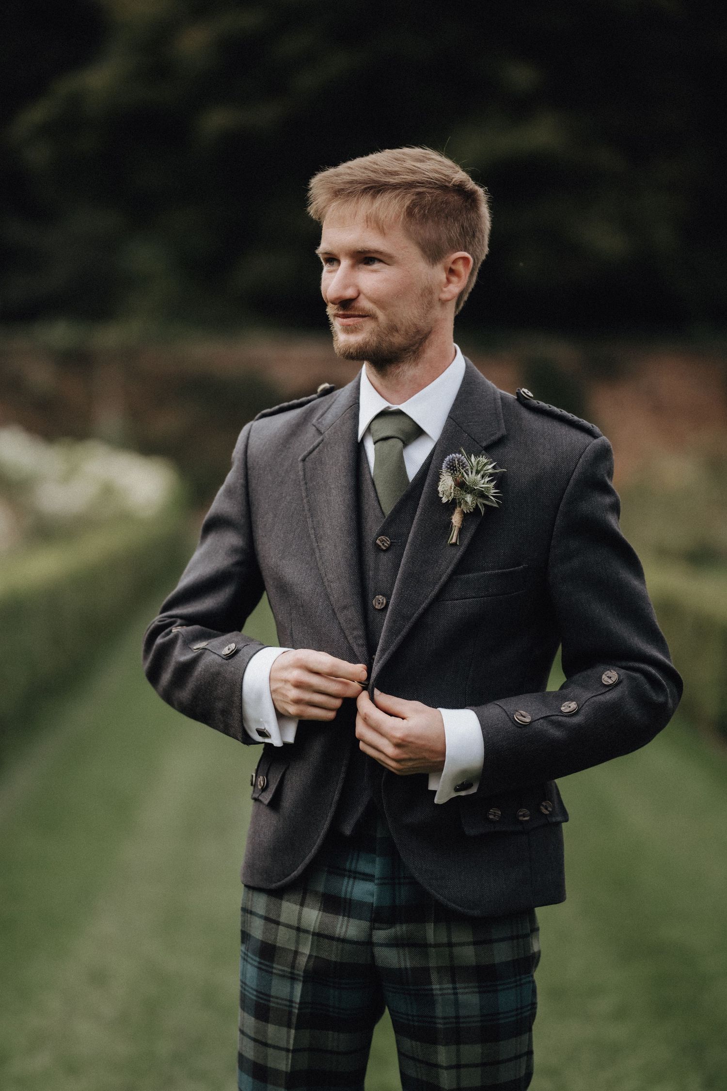 A person in a formal grey suit and tartan kilt adjusting their jacket outdoors.