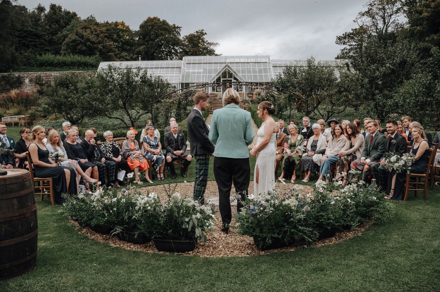 Outdoor garden wedding ceremony with guests seated around a circular floral arrangement.