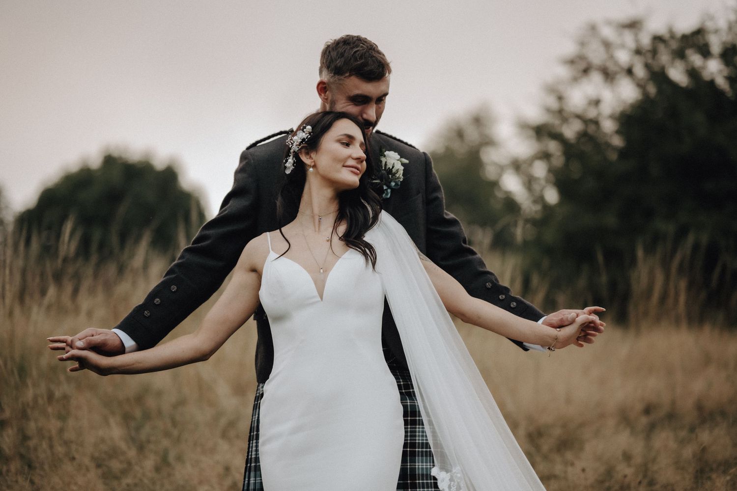 Newlywed couple poses romantically in a field with bride's veil flowing in the breeze.