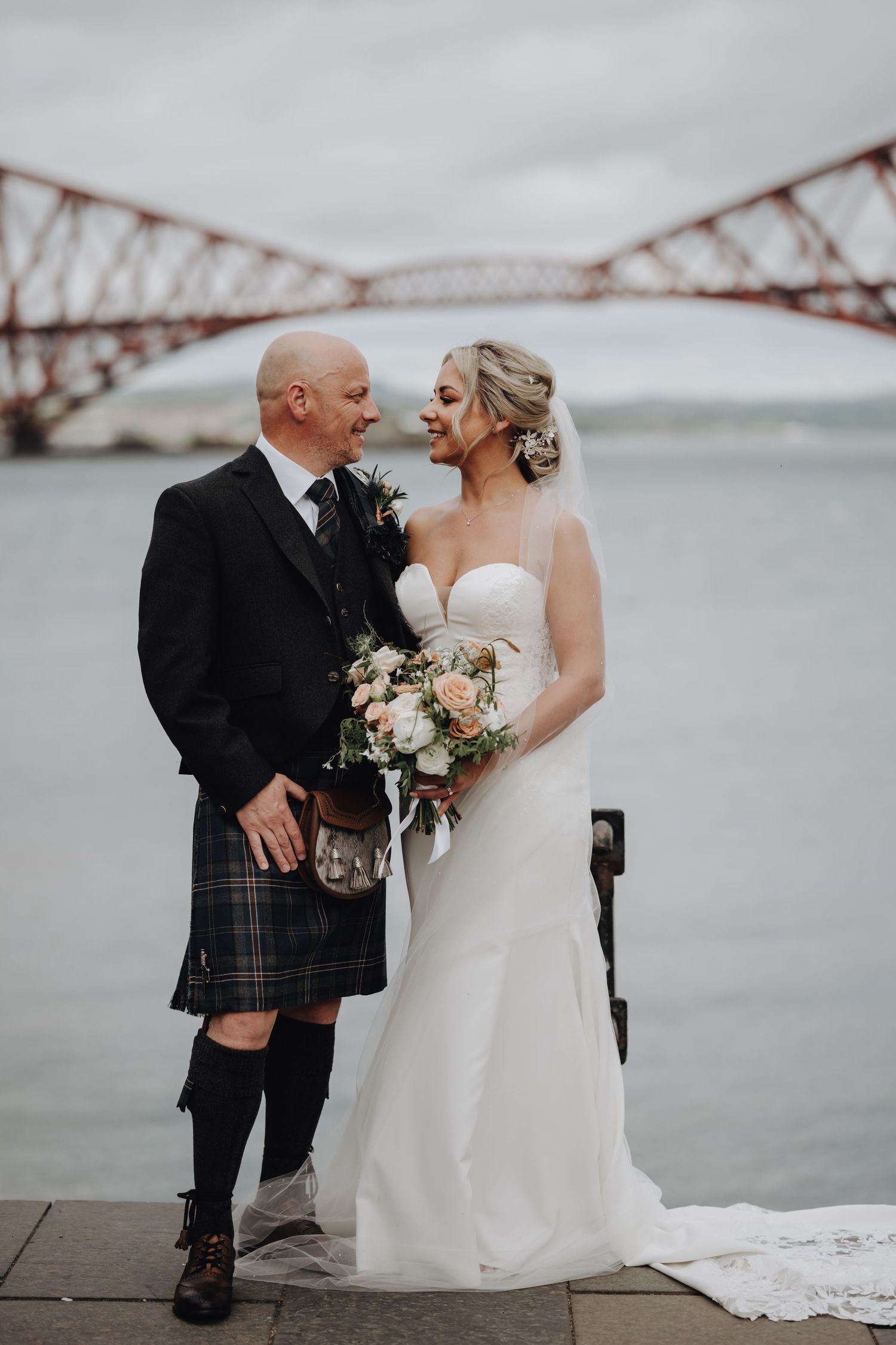 A wedding couple poses against iconic Forth Bridge in Scotland with bride in white dress and groom in traditional kilt.