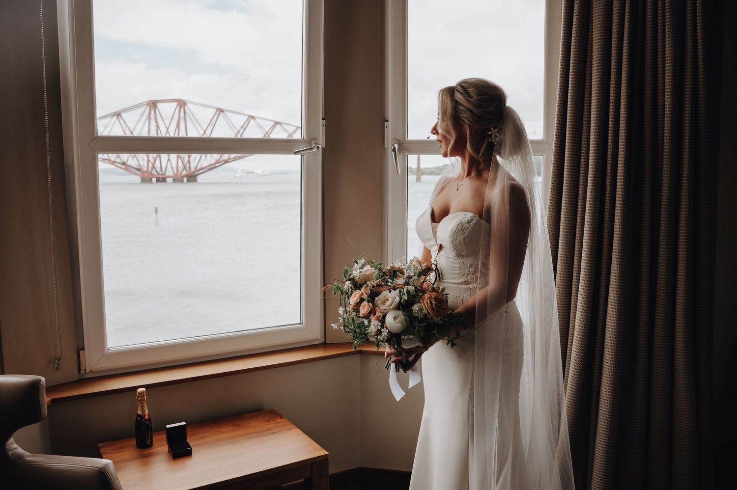 Bride holding bouquet gazes out window with bridge visible in background.